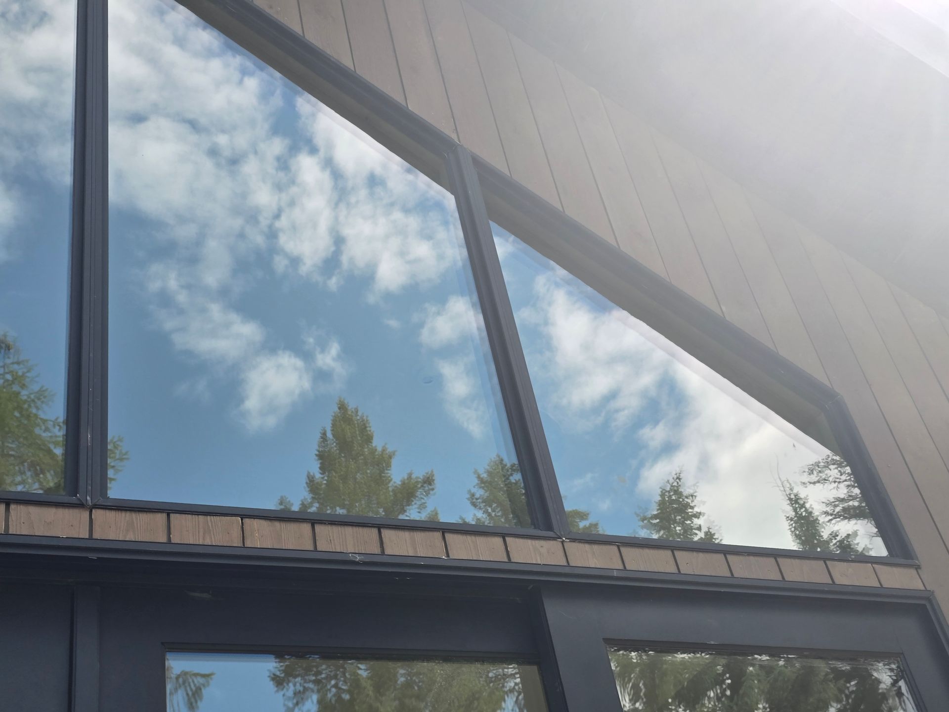 A slanted building roofline with dark window frames reflecting a bright, cloudy blue sky and green pine trees.