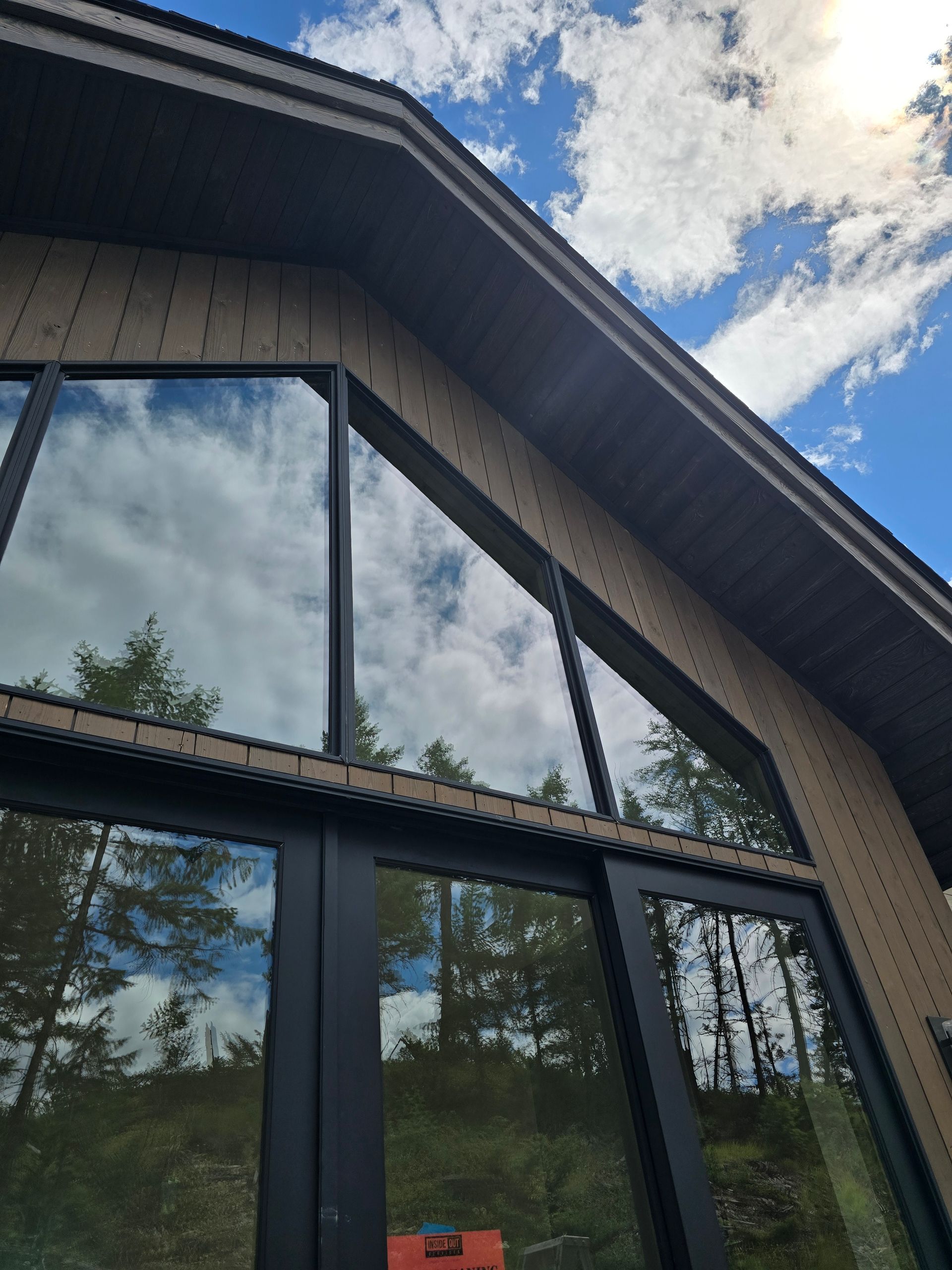 A low-angle view of a building's gabled wooden exterior with large, reflective windows mirroring trees and a blue sky.