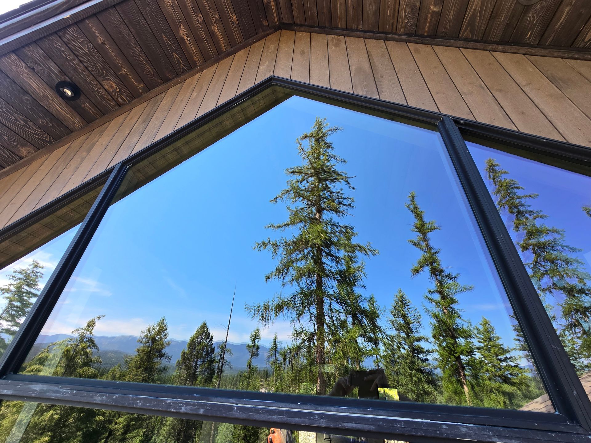 A triangular window reflects a sunny forest landscape and blue sky against a wooden exterior wall.