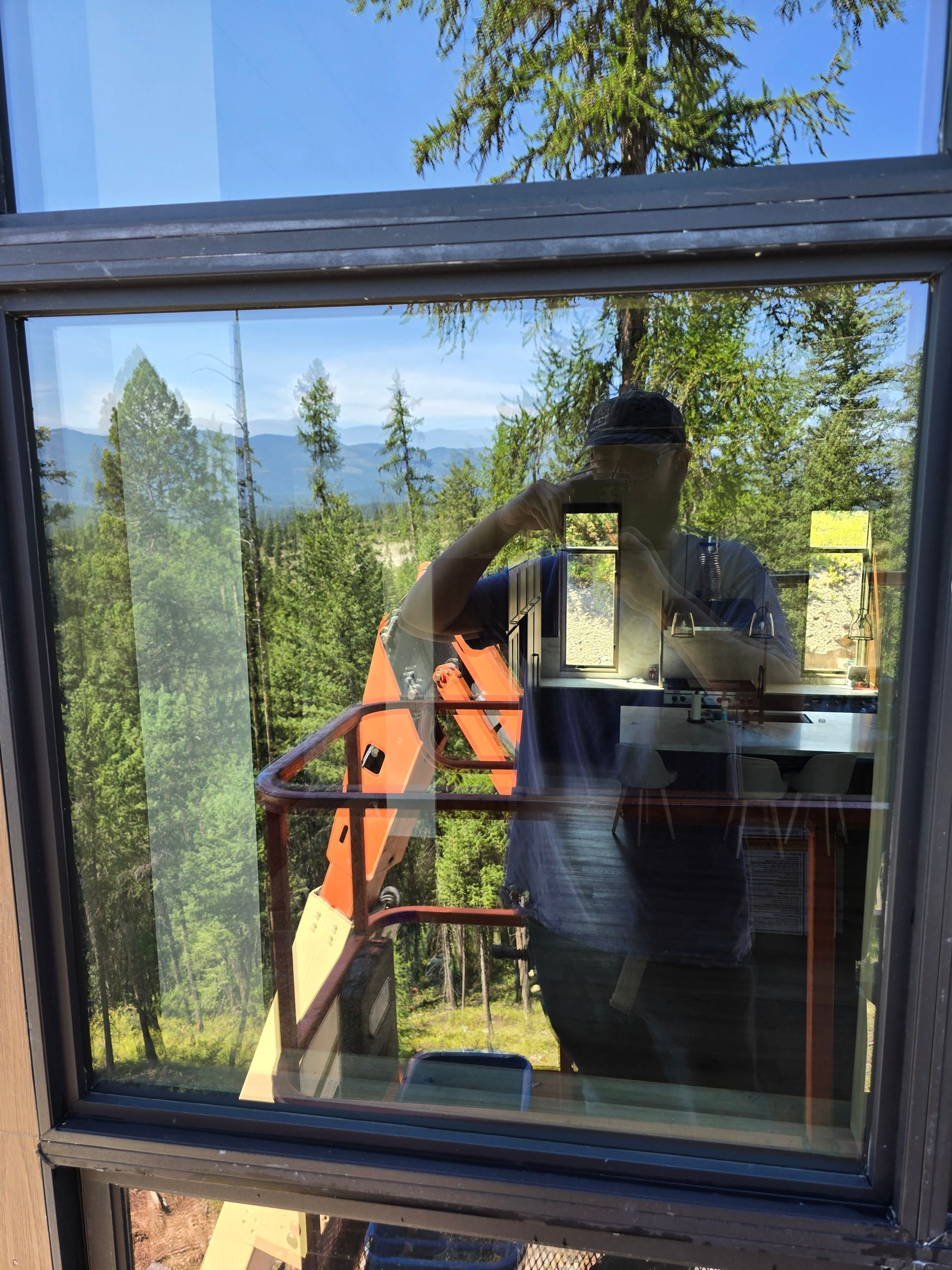 A person taking a selfie in a window reflection, revealing an orange construction lift outdoors against a forest backdrop.