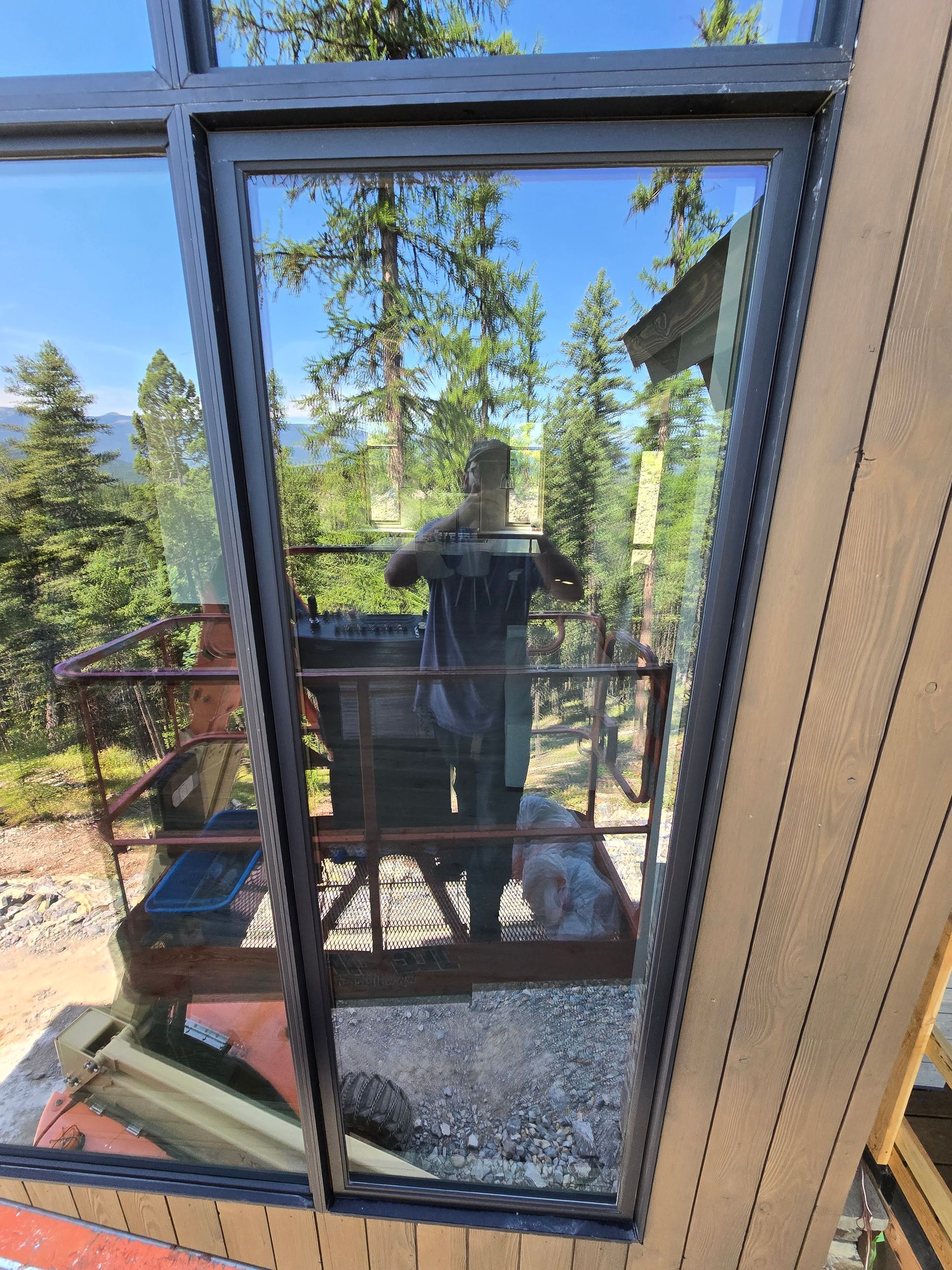 Reflection of a person holding a phone, visible through a large window frame overlooking a wooded, mountain landscape.