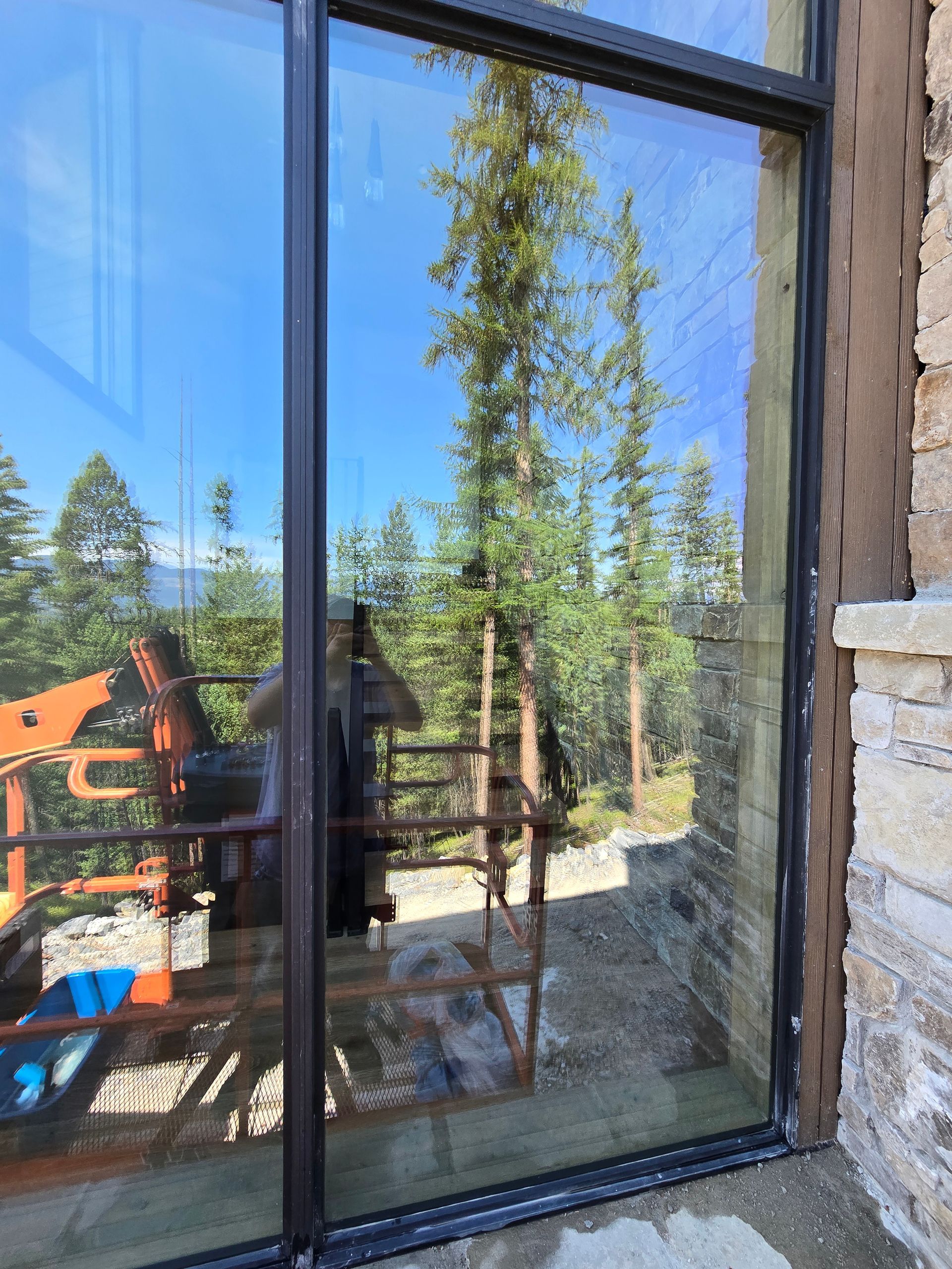 A glass door reflects a forest of pine trees, a bright blue sky, and construction equipment outside a stone building.