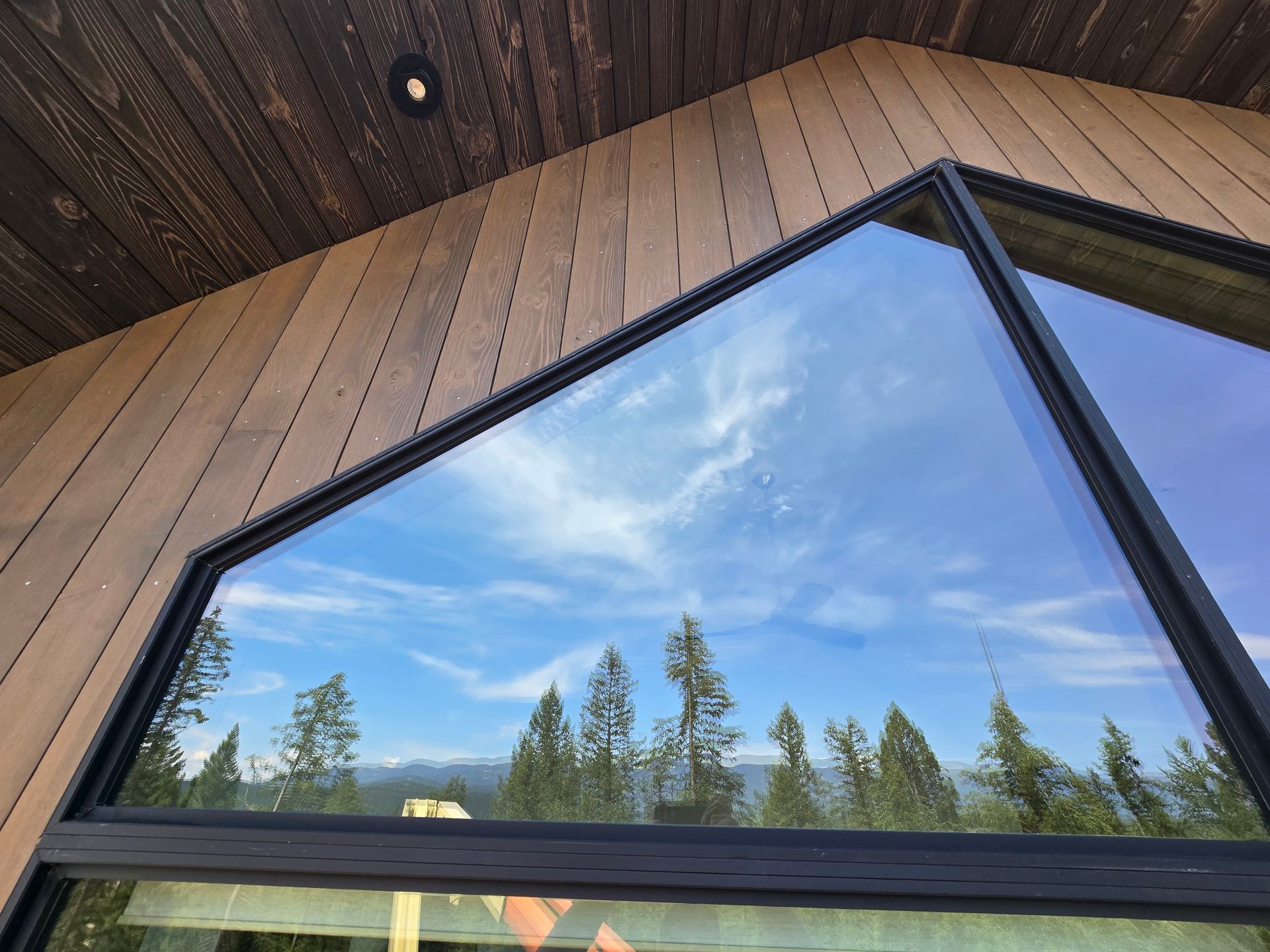 A view of a blue sky and pine trees reflected in a large, modern, black-framed angled window on a wood-paneled house.