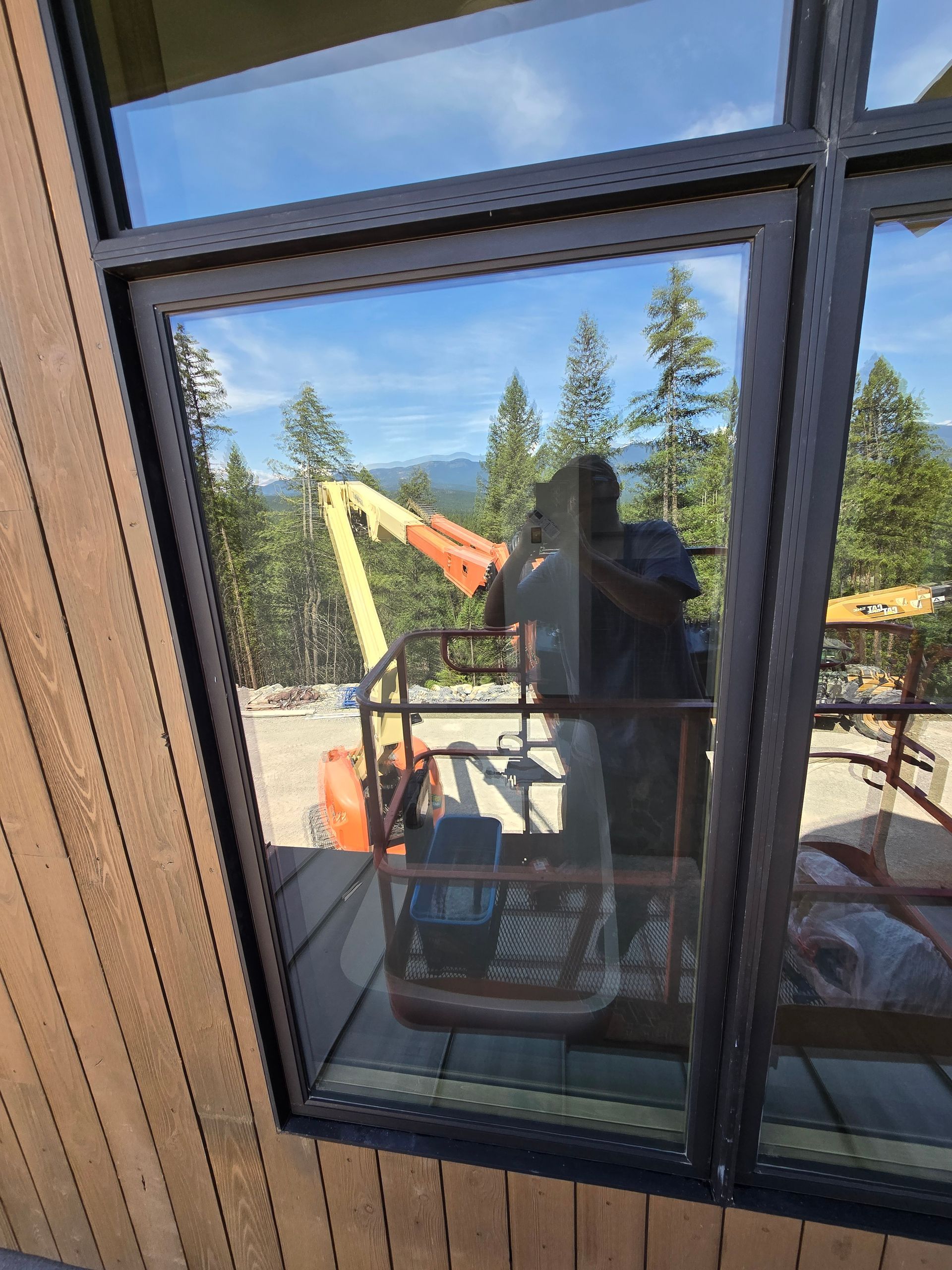 A window reflection showing a person operating an orange aerial lift outdoors near trees.