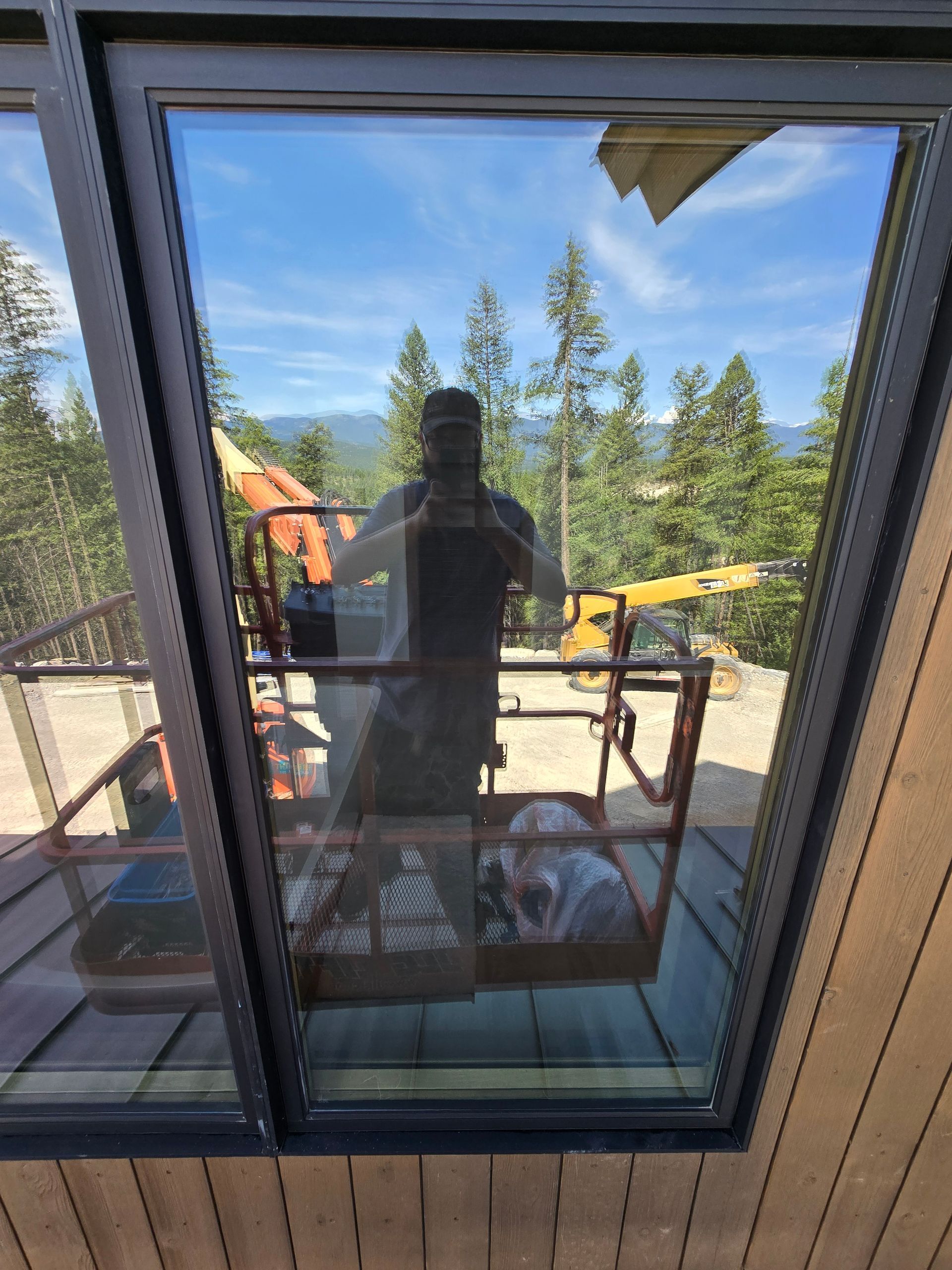 A reflection in a window shows a person standing on a mobile elevating work platform against a backdrop of pine trees.