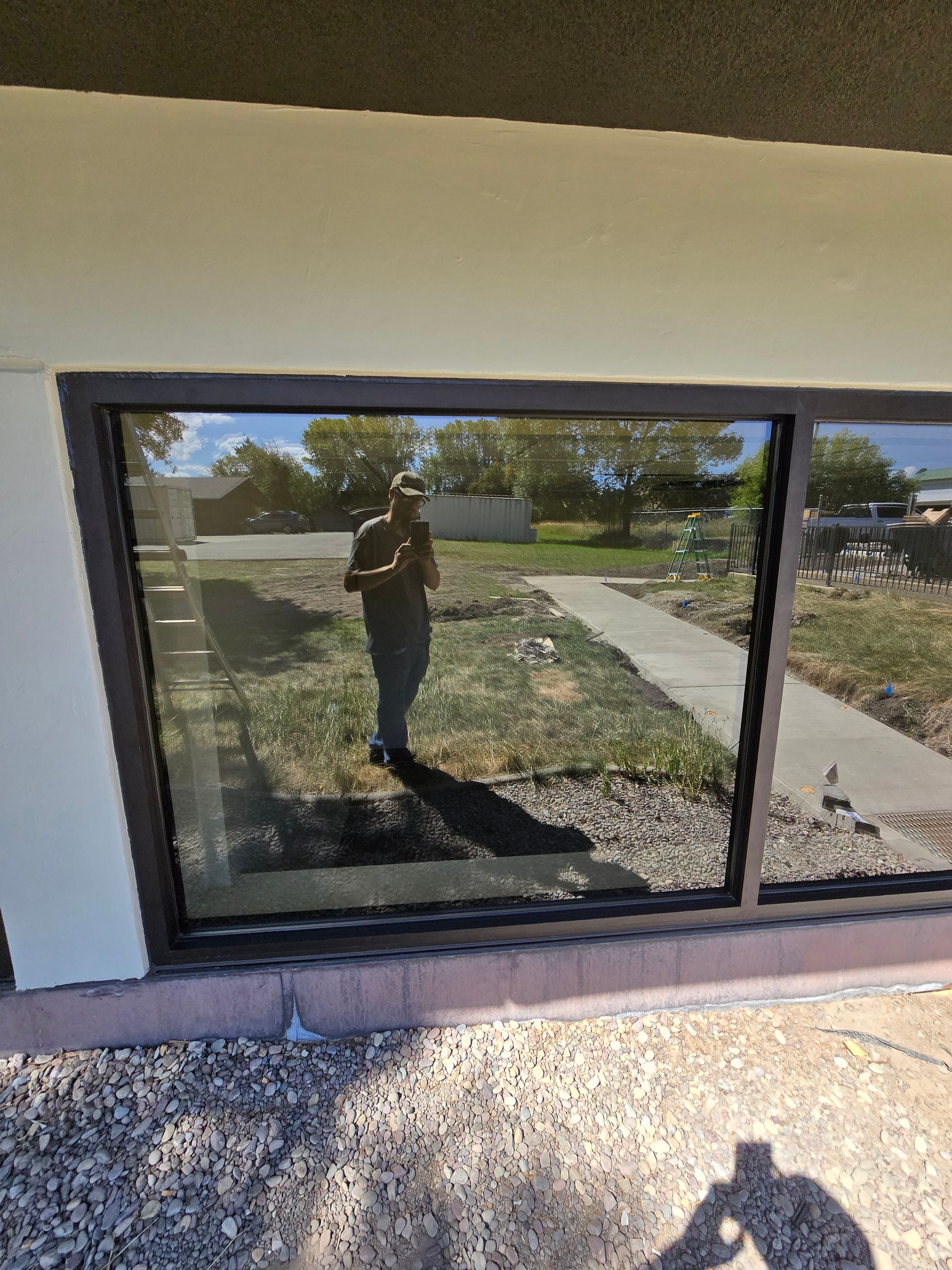 A person stands in front of a dark-framed glass window reflecting a yard, sidewalk, and trees.
