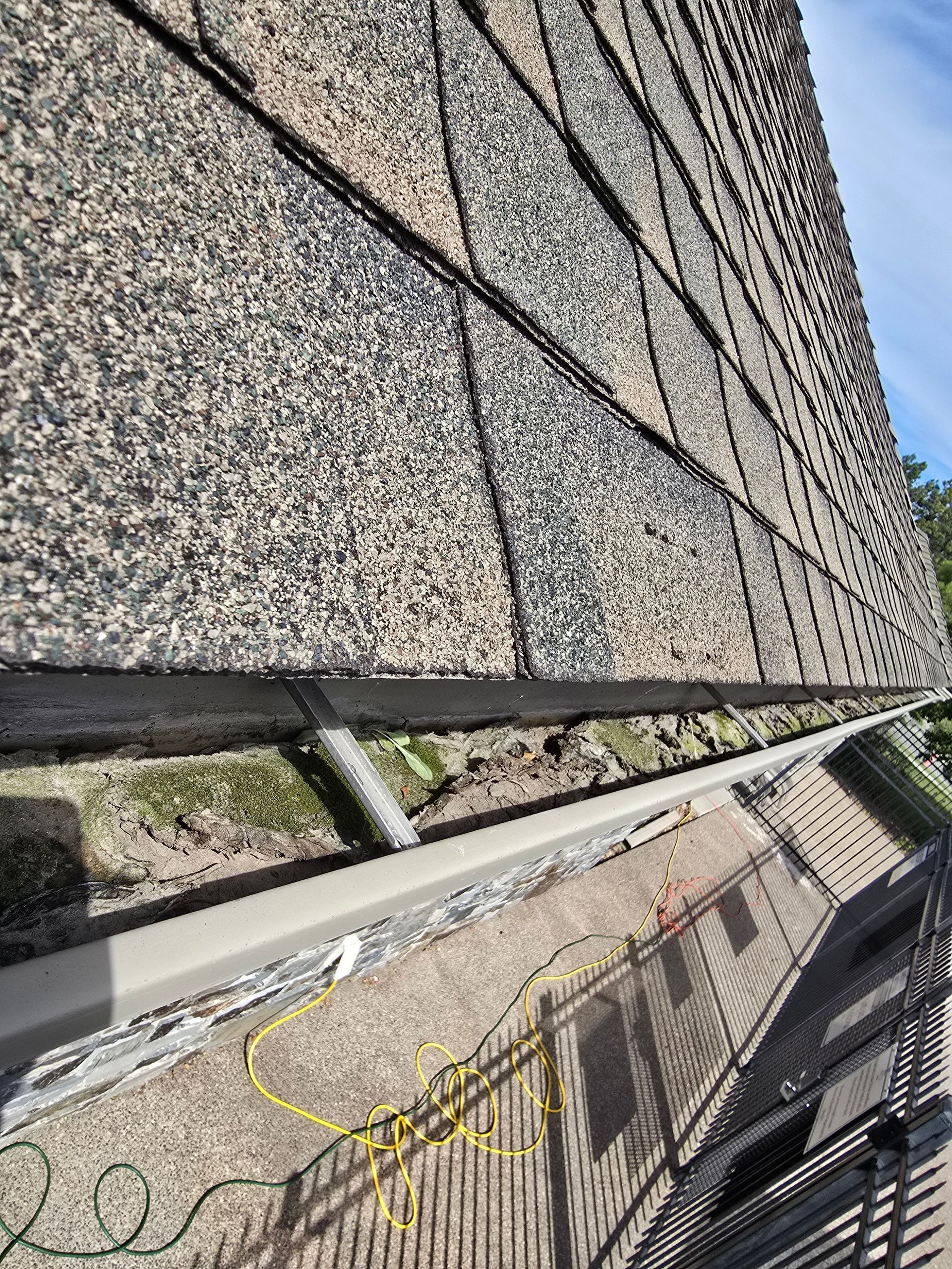 An angled view of a roof edge with clogged gutters, with a yellow cord and roof debris visible on the ground below.