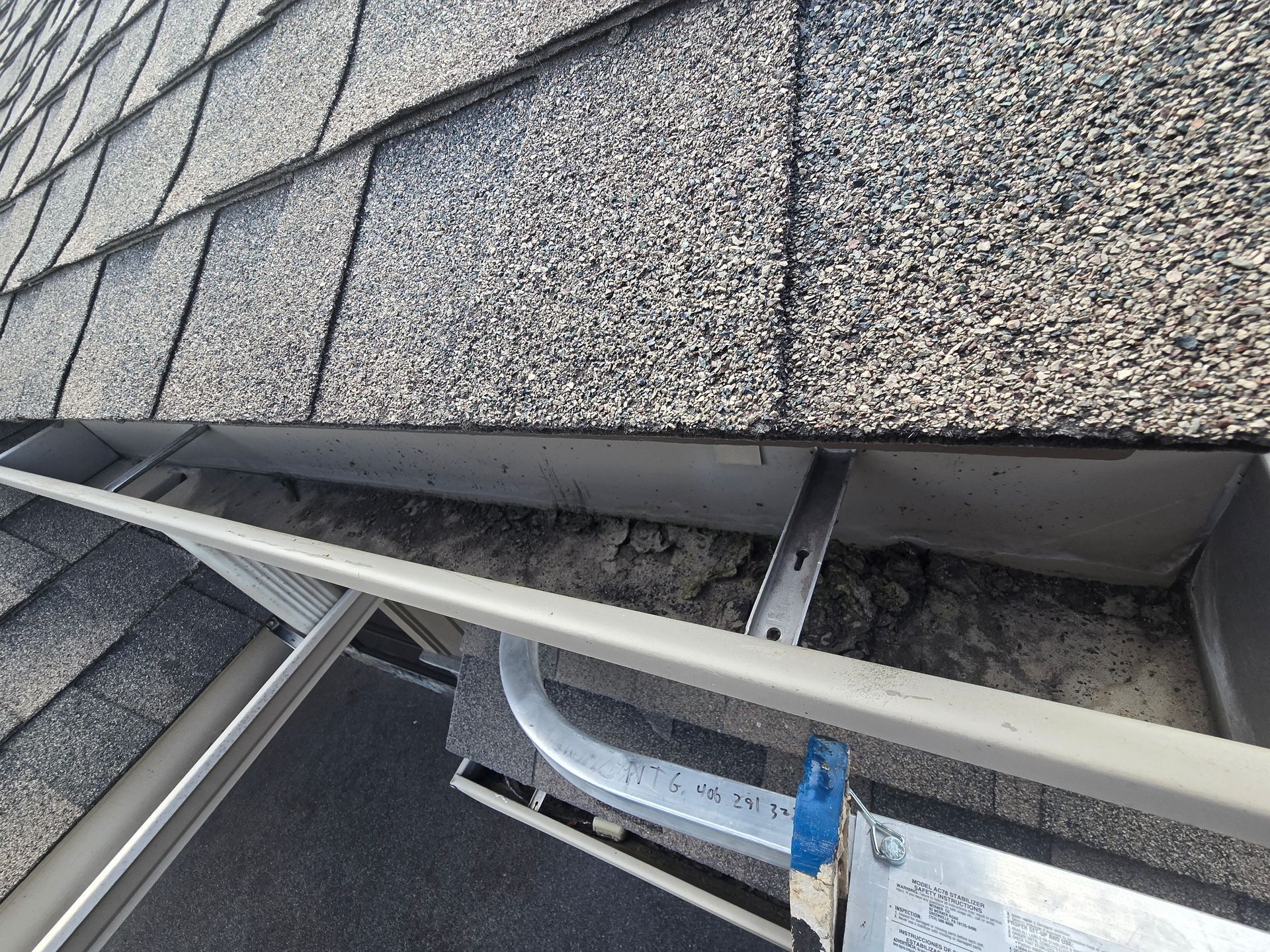 A close-up view of a metal roof gutter filled with debris, showing a metal hanger bracket and a ladder rung below.