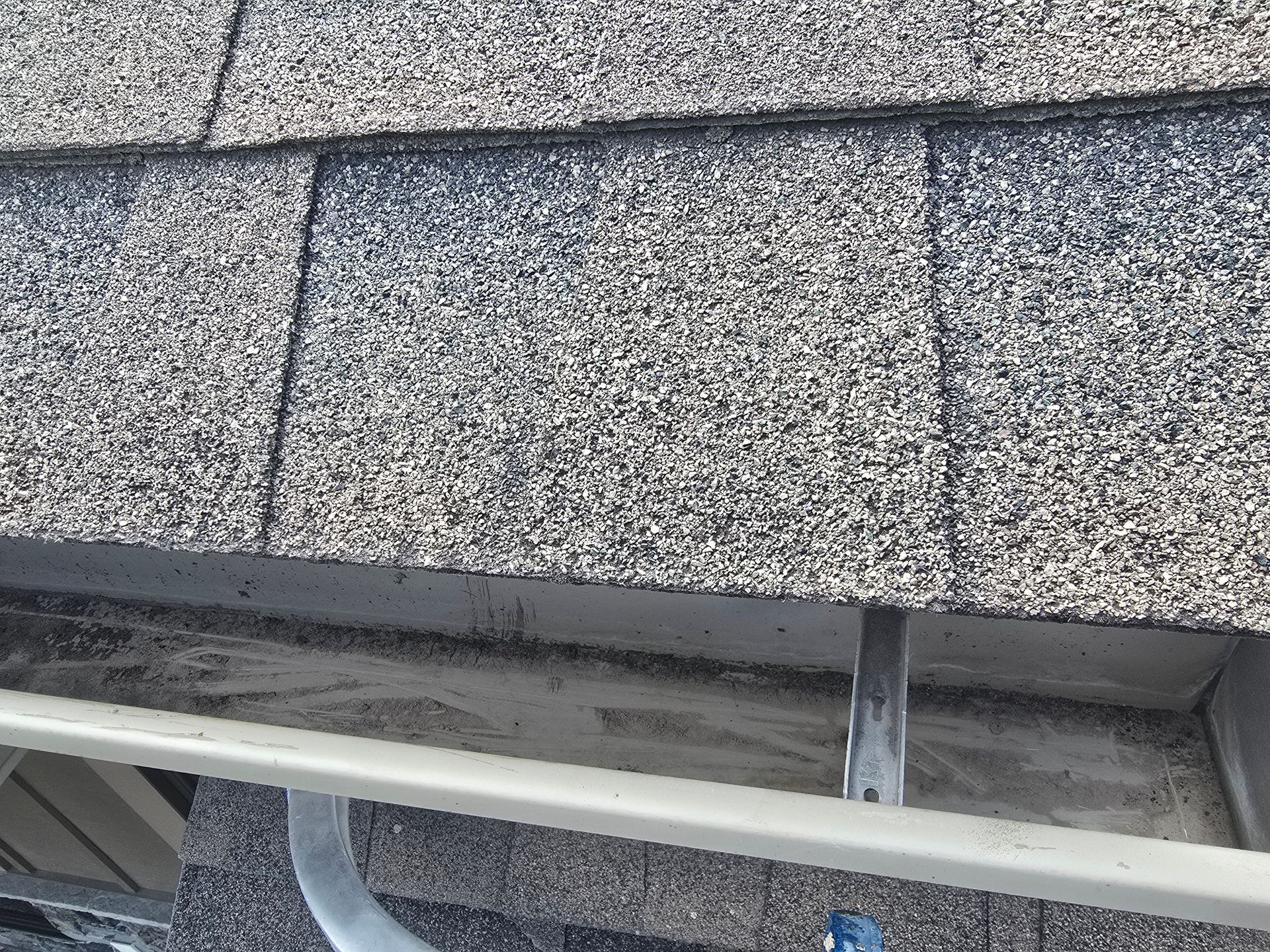 A close-up view of gray asphalt roof shingles hanging over a beige rain gutter.