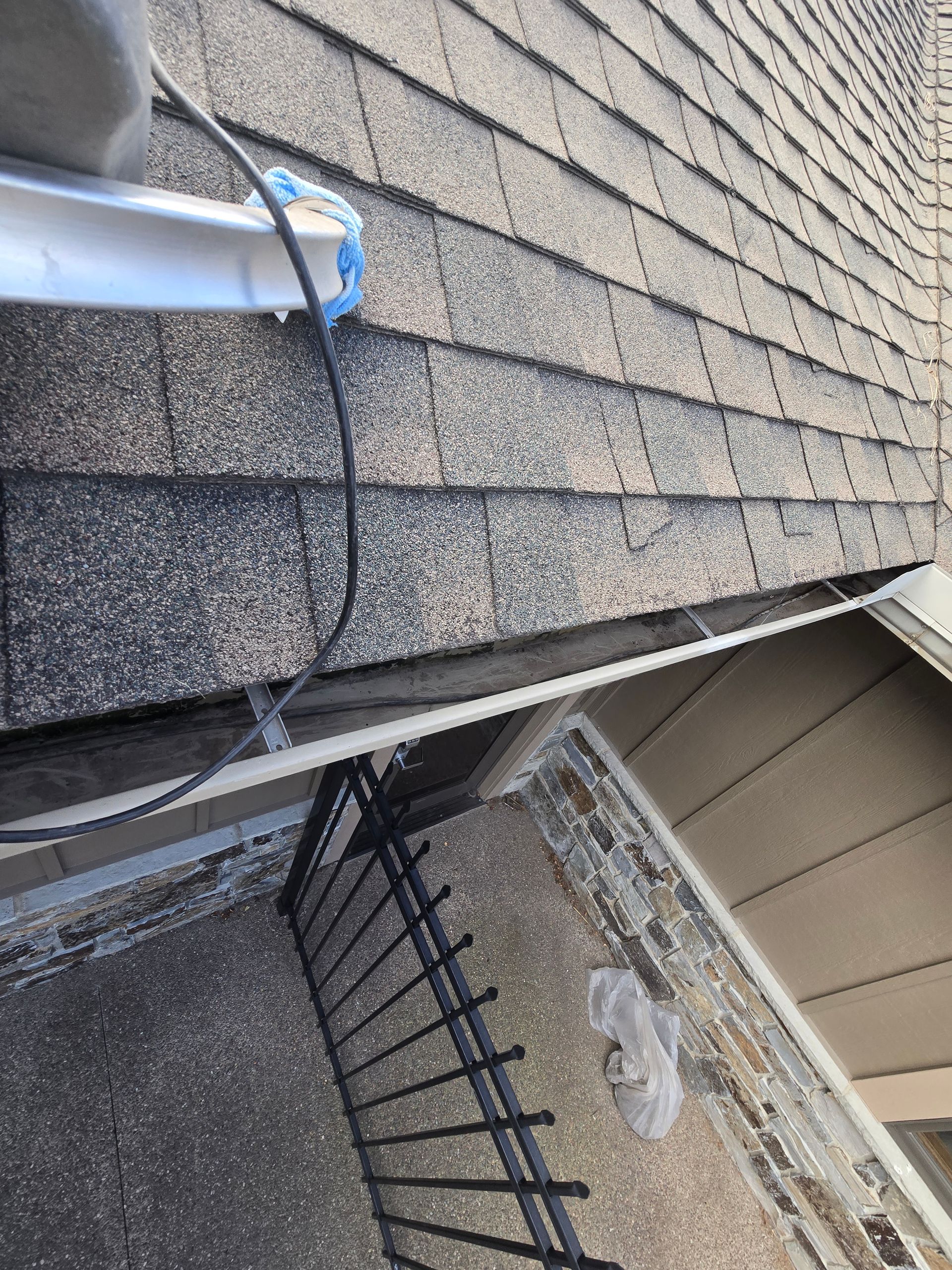 A view from above showing a dark asphalt shingled roof, a gutter, and a metal railing leading down to a stone walkway.