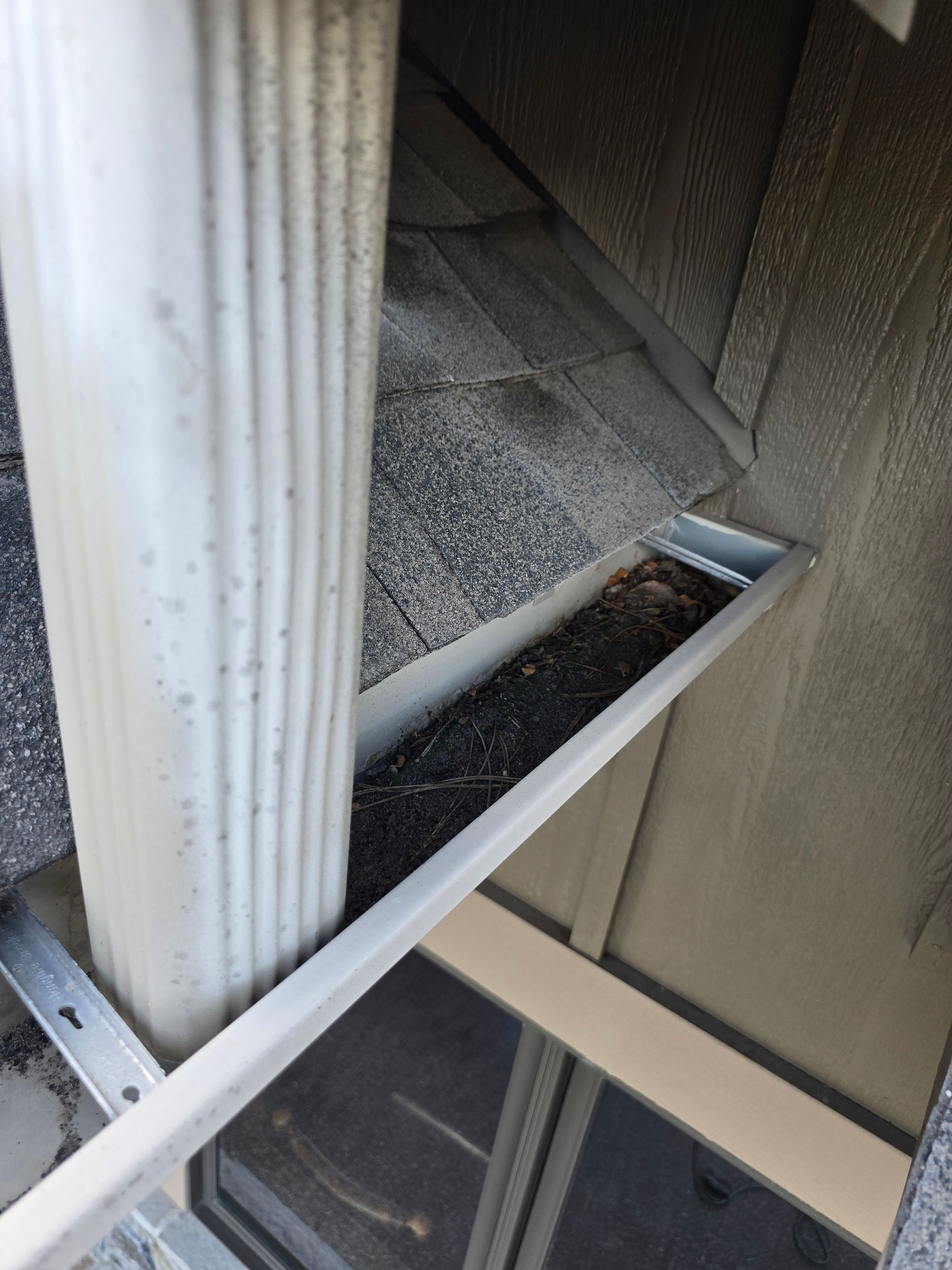 A metal gutter overflowing with dark debris and sediment, situated below a white downspout and shingled roof.