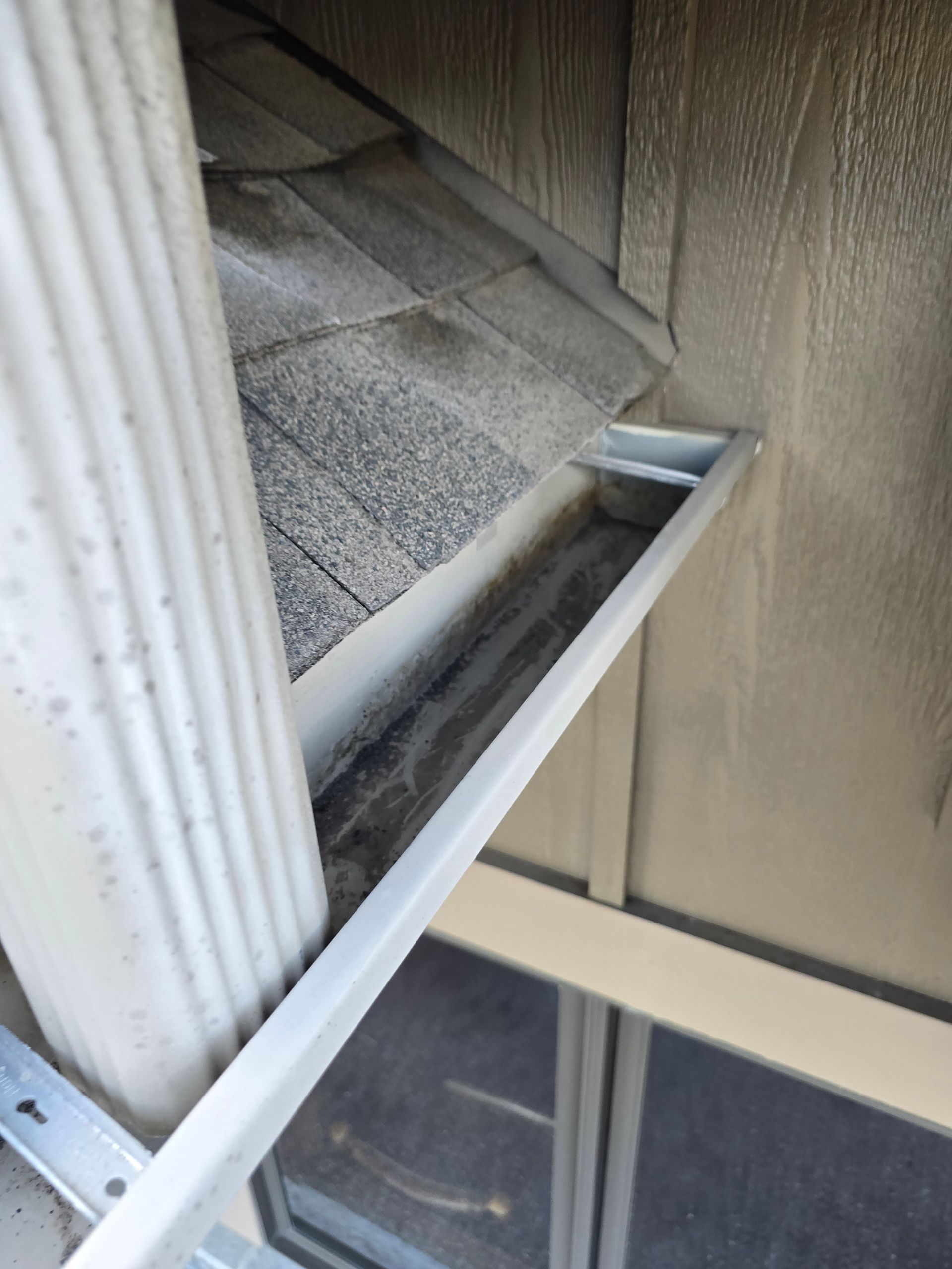 A white metal downspout runs vertically next to a tan house wall, positioned over a partially visible roof gutter.