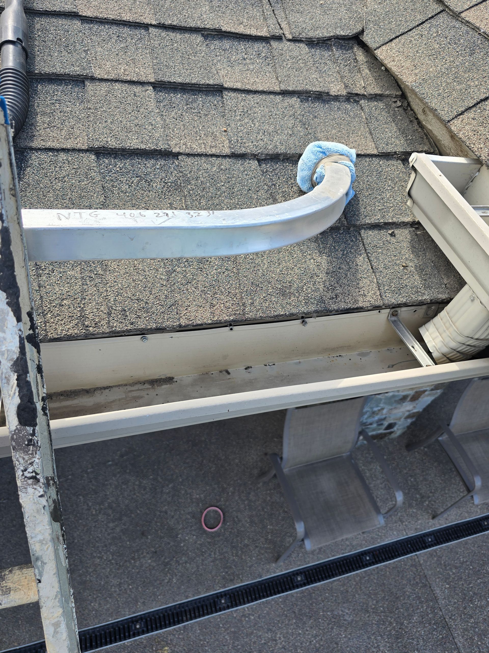 A metal pipe with a blue foam cover rests on an asphalt shingle roof above a partially installed gutter section.