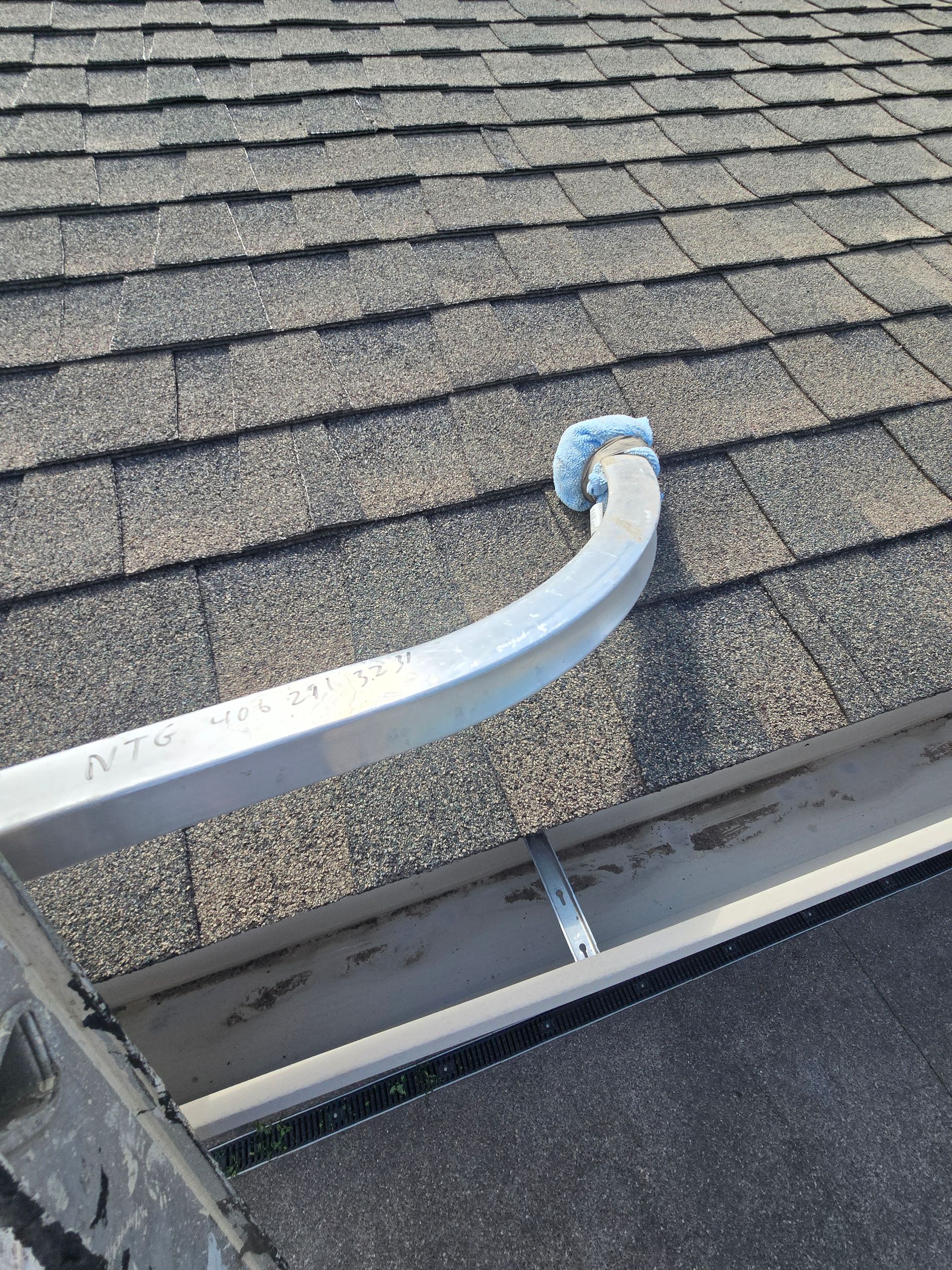 A curved white gutter extension rests on a shingled roof, leading into a metal gutter section secured by a strap.