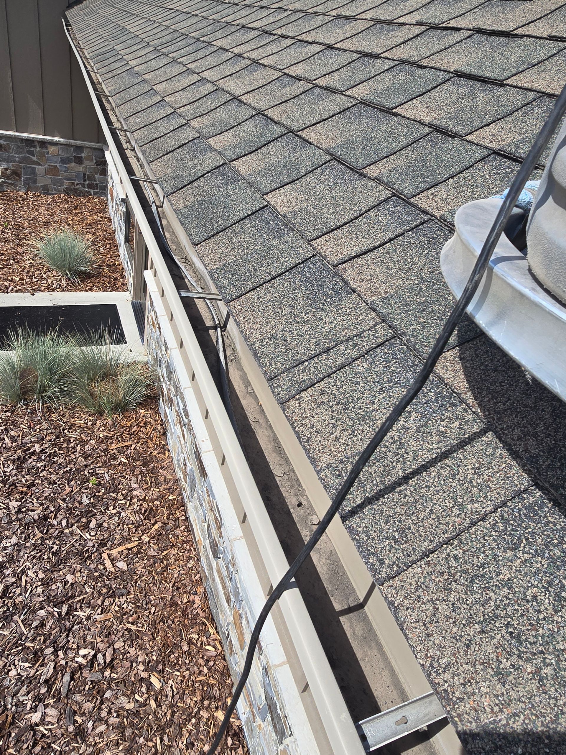 A black power cord hangs across a tan gutter along the edge of a gray shingled roof above a gravel yard.