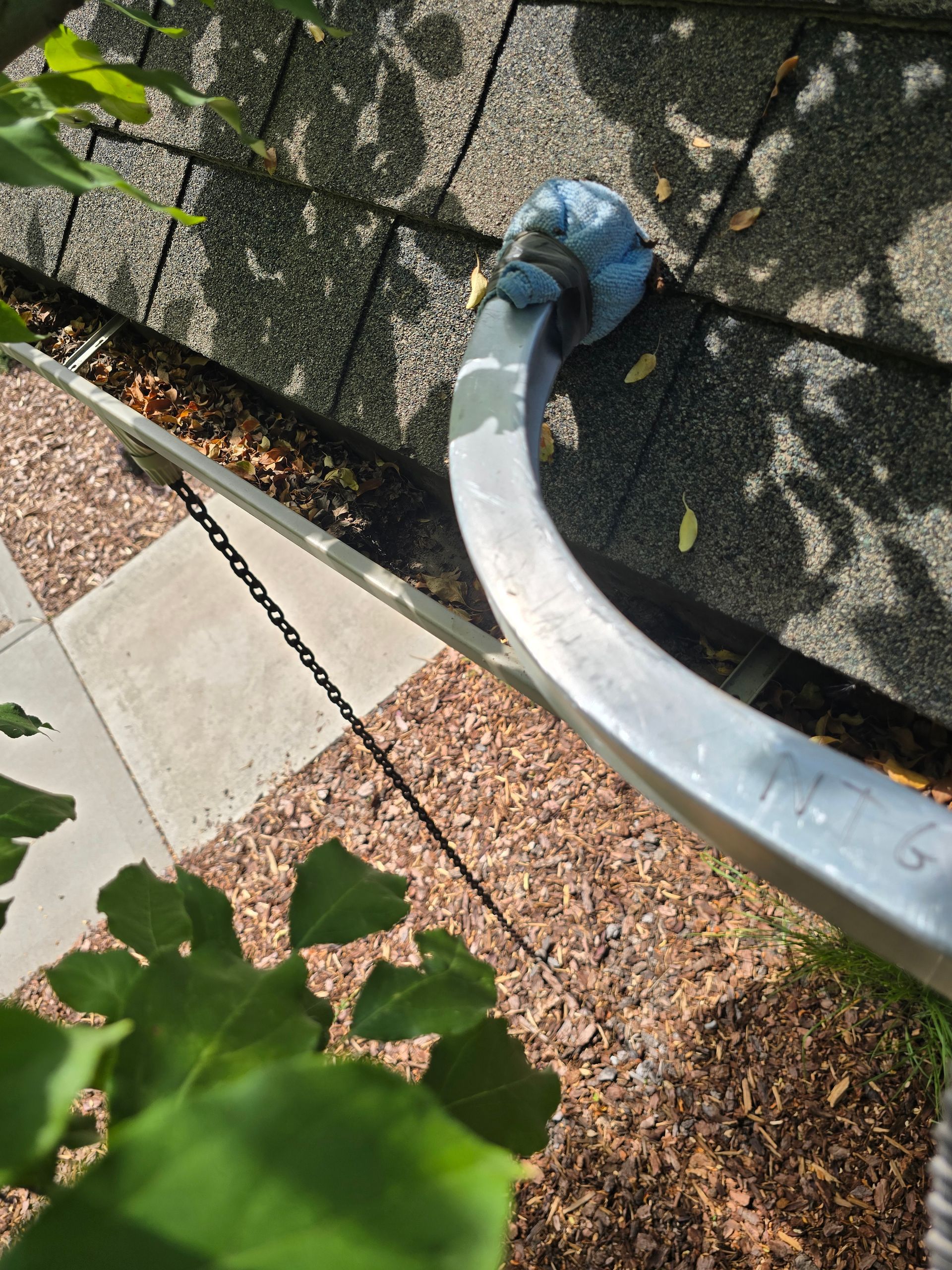 A curved metal pipe emerges from a shingled roof, next to a rain gutter filled with dry leaves and a hanging metal chain.