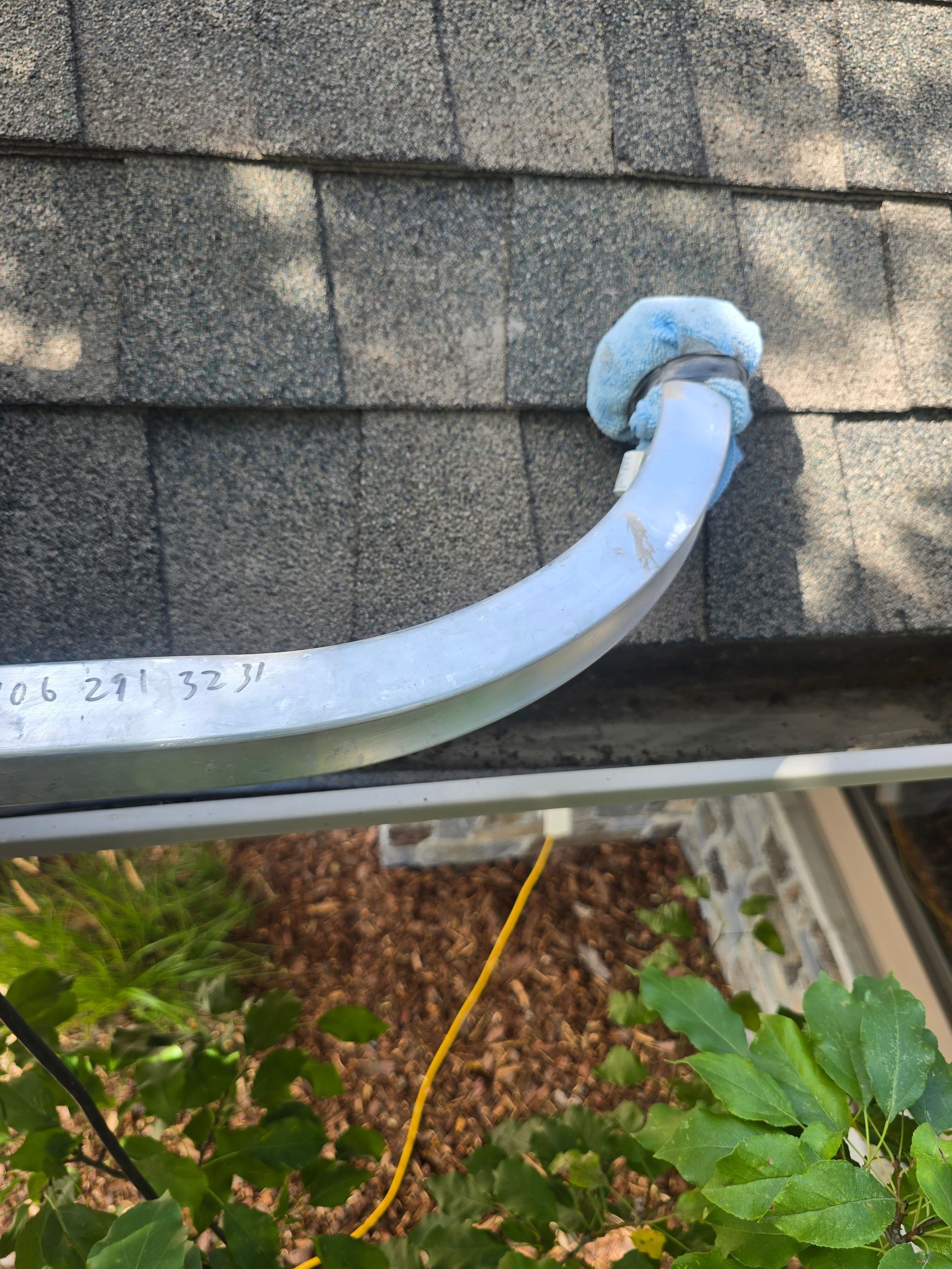 A metal ladder stabilizer with a light blue foam pad resting on the corner of a shingled roof above a gutter.