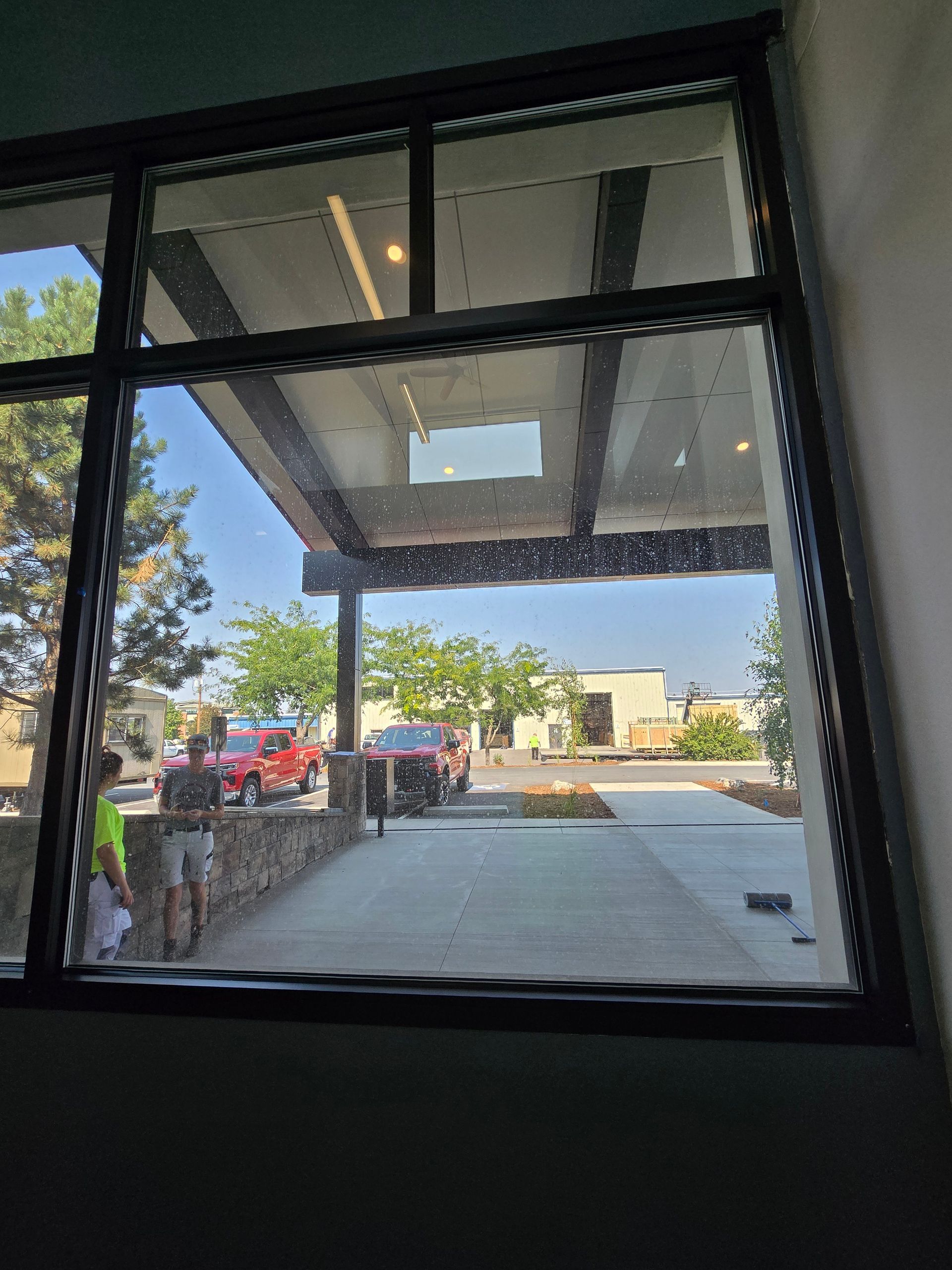A view through a window showing a bright outdoor area with a sidewalk, parked vehicles, trees, and people standing.