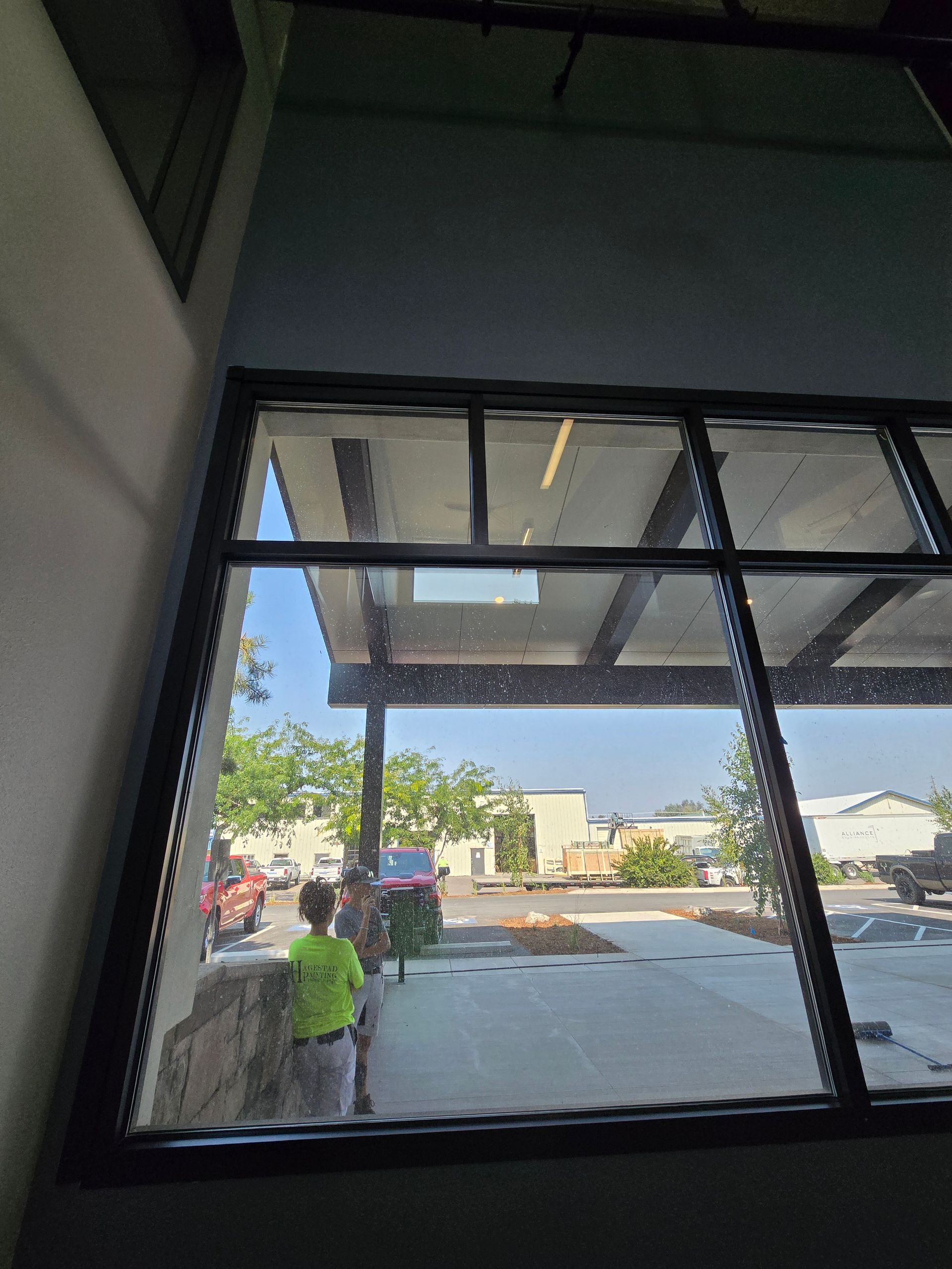 A person in a bright green shirt stands outside a large window looking into a building on a sunny day.