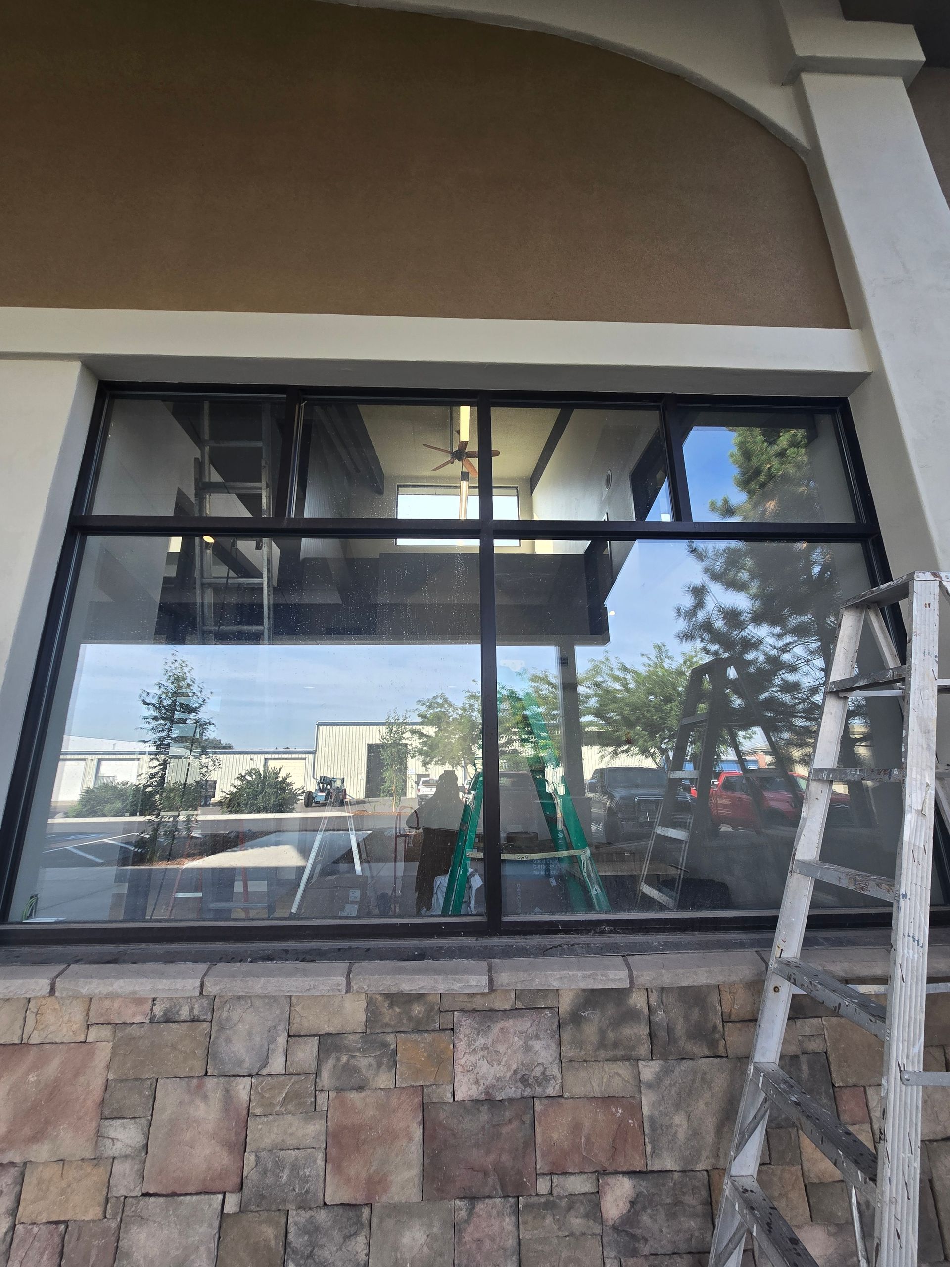 A large store window with a black frame reflects an outdoor parking lot, with a ladder leaning against the stone base.
