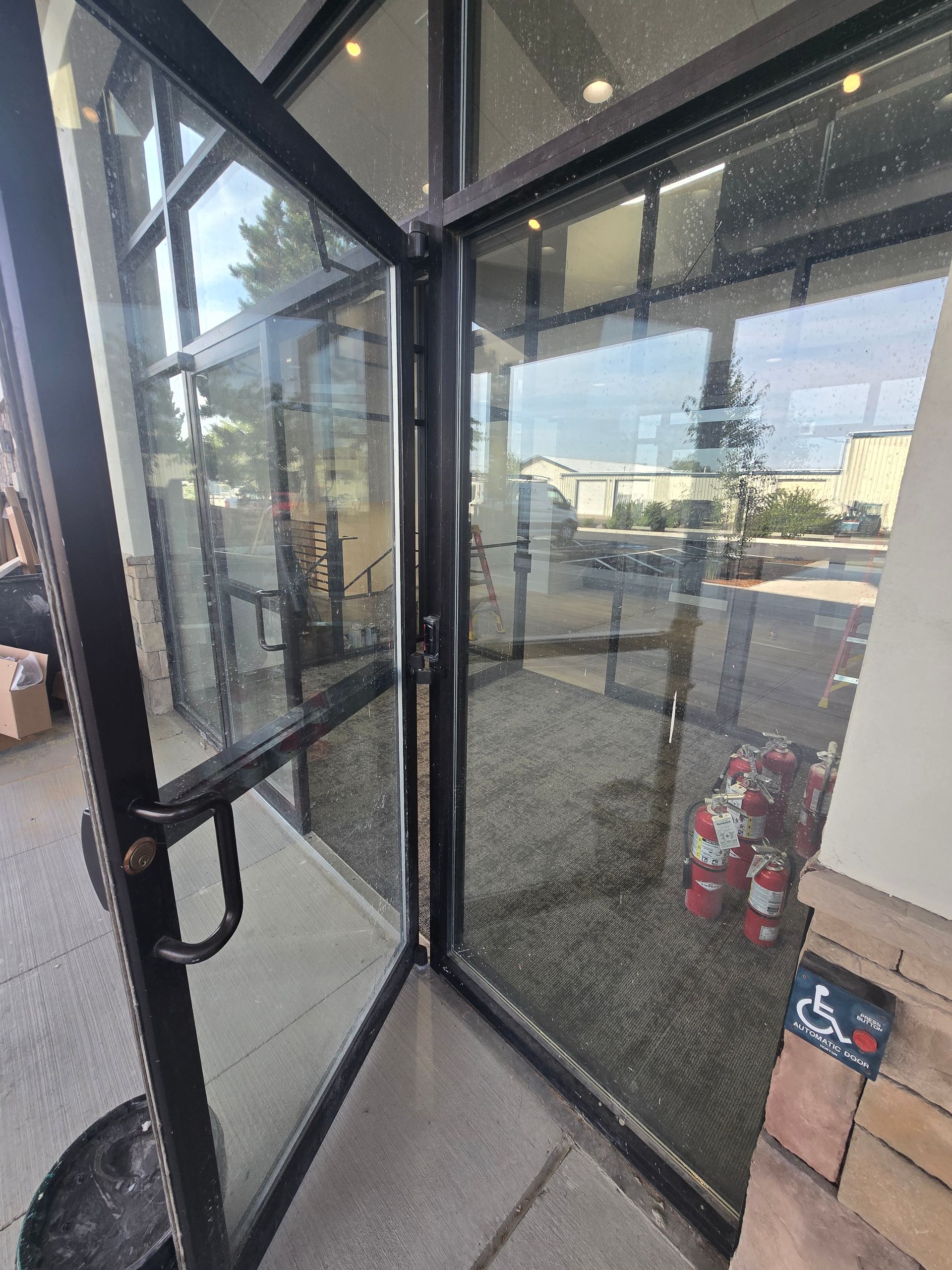A glass storefront entrance with a dark metal frame, showing a paved walkway and building exterior through the reflection.