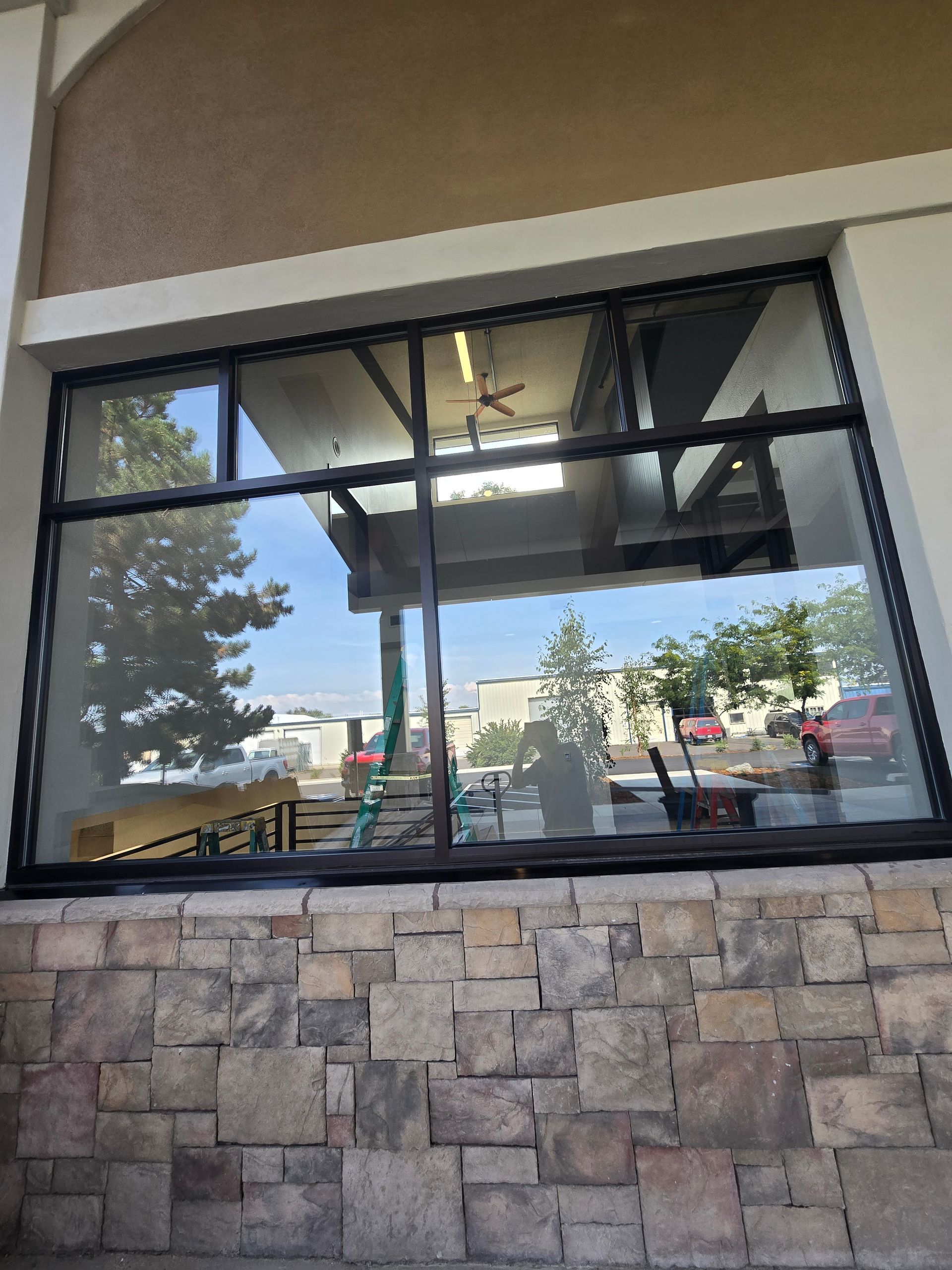 A large storefront window with a black frame, reflecting a parking lot and trees above a stone-tiled base.