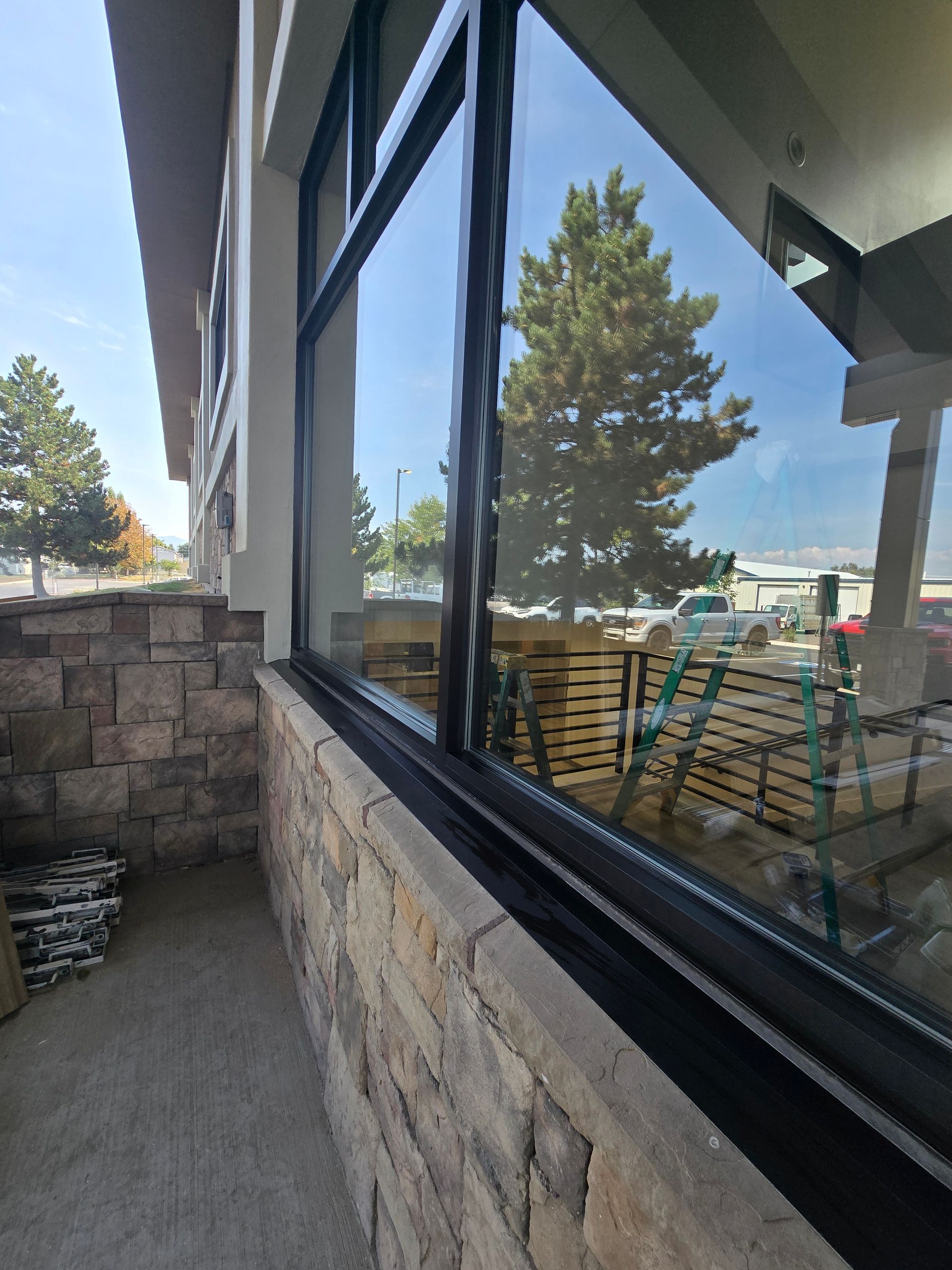 A side-angle view of a stone wall base and a tall glass storefront window reflecting trees, a parking lot, and sky.