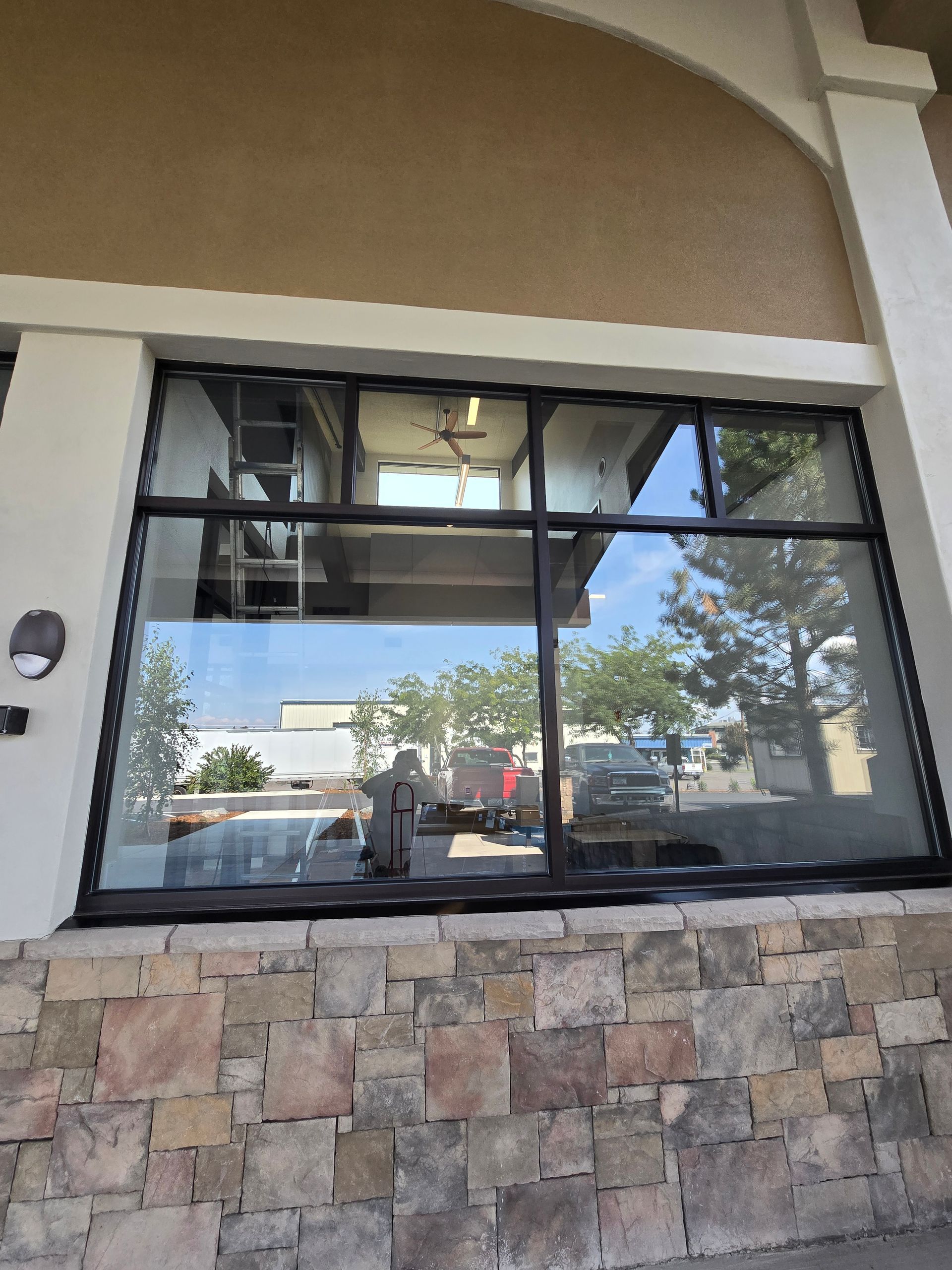 A large rectangular storefront window with a black frame, reflecting a sunny outdoor parking area and nearby trees.