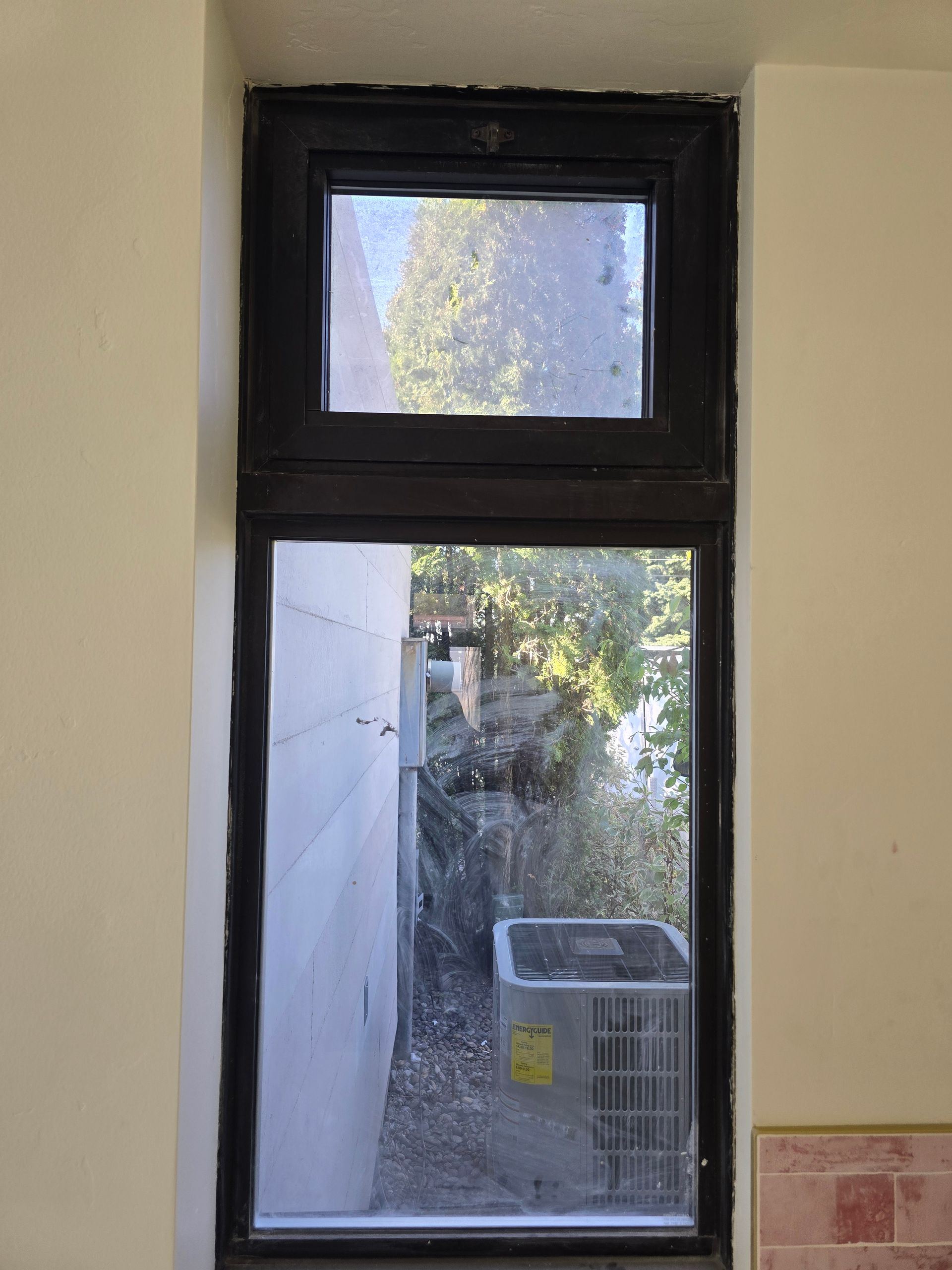 A view through a dark-framed, multi-pane window looking out at a side yard with an outdoor air conditioning unit.