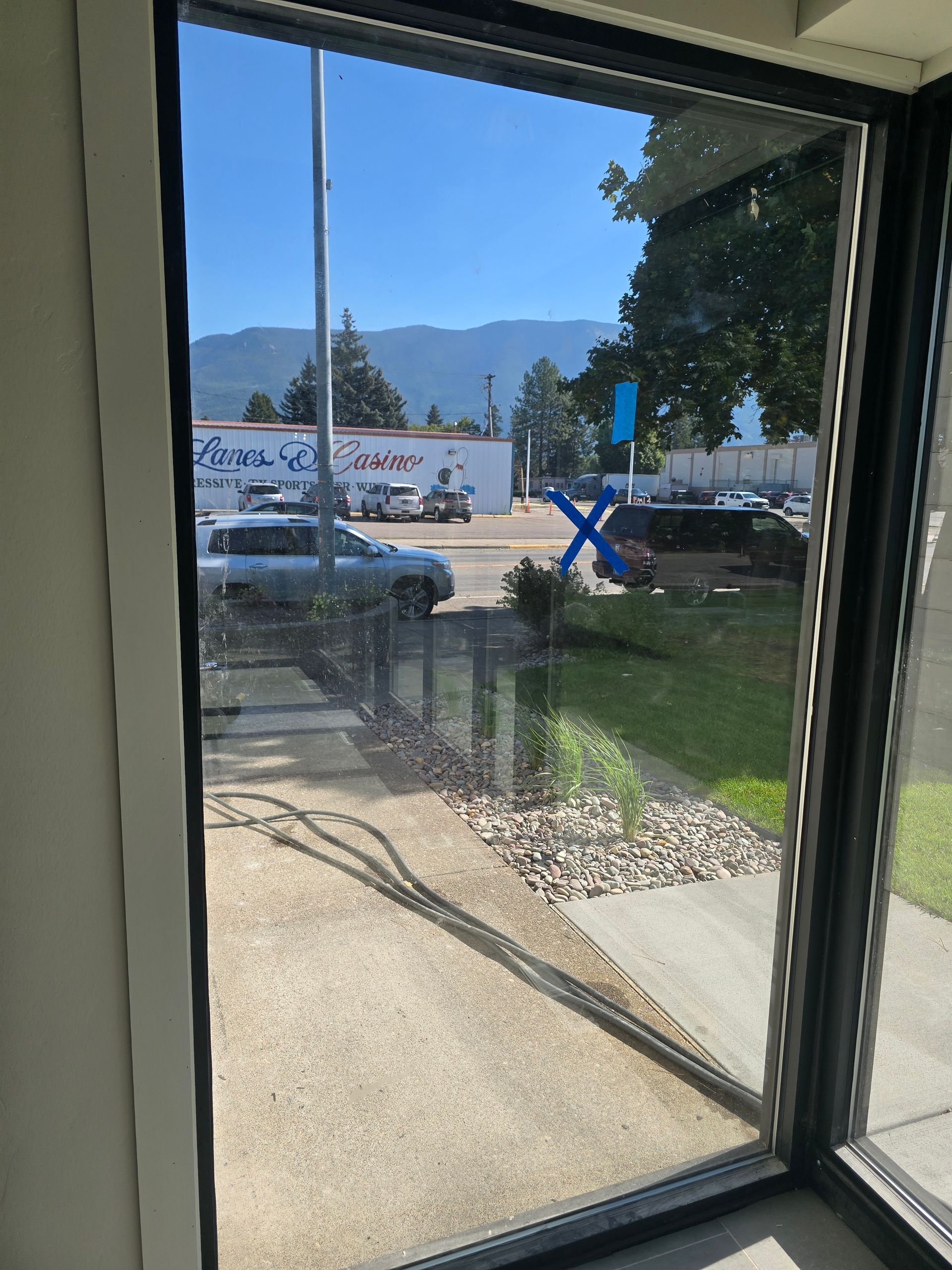 A view through a window looking at a parking lot, a white building with text, trees, mountains, and a blue X on the glass.