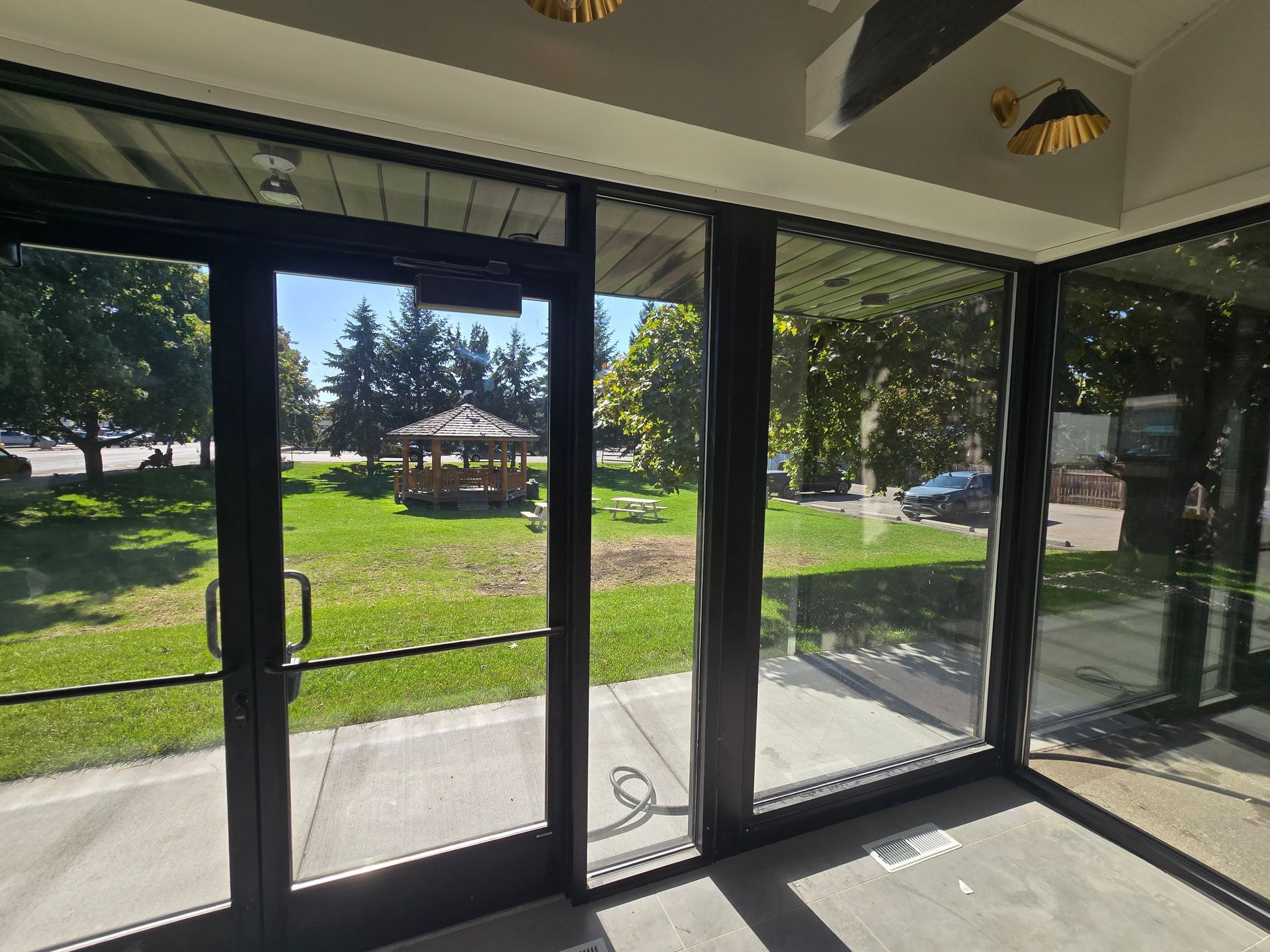 A view through glass double doors and floor-to-ceiling windows showing a sunny park with a wooden gazebo and lawn.