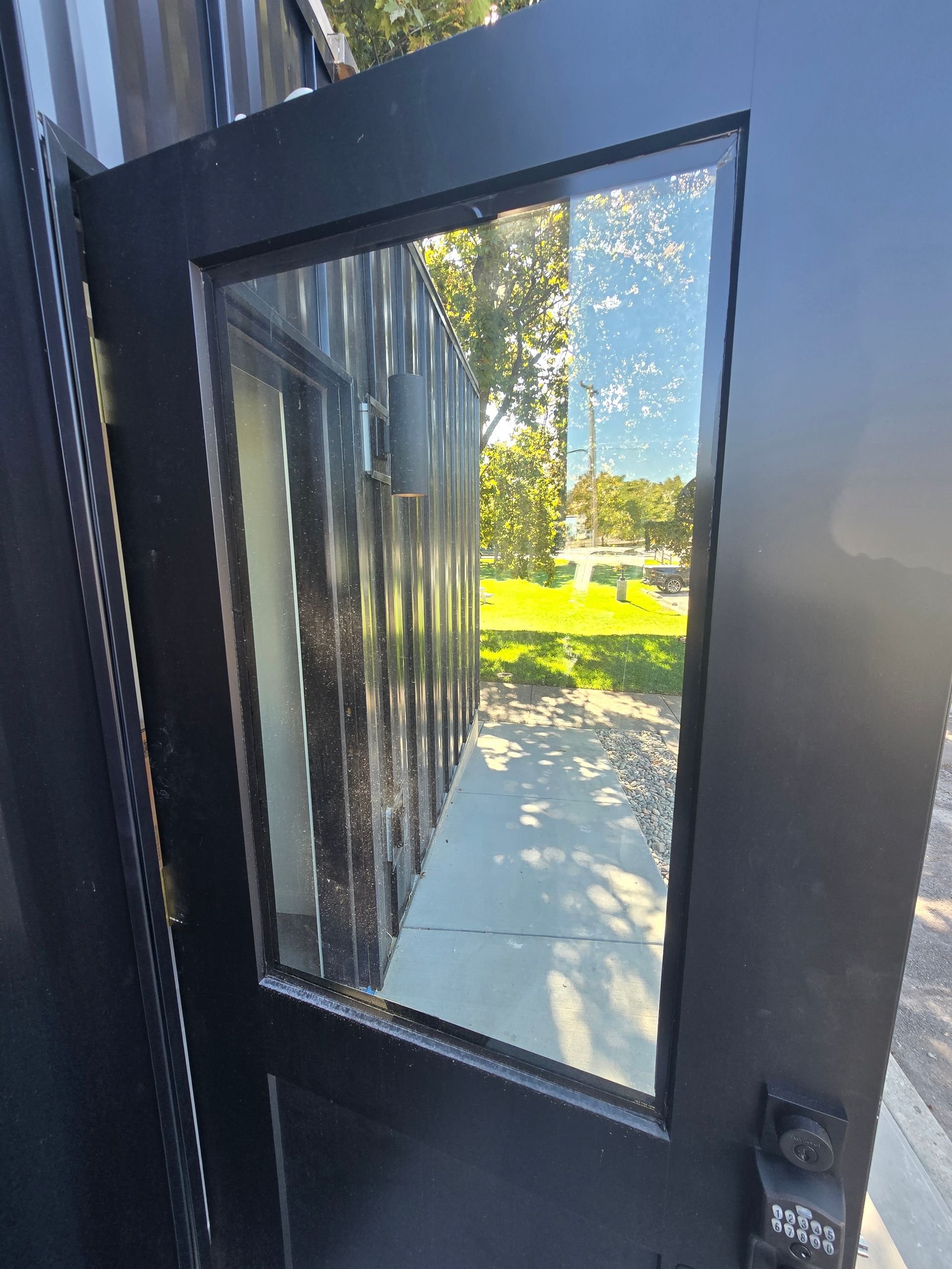 A black door with a large glass window pane, reflecting a building exterior, trees, and grass in the sunlight.