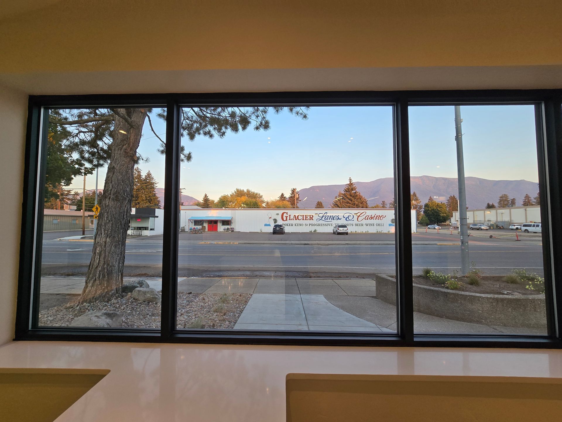 View through a large rectangular window showing a tree, a street, a building with signage, and distant mountains.