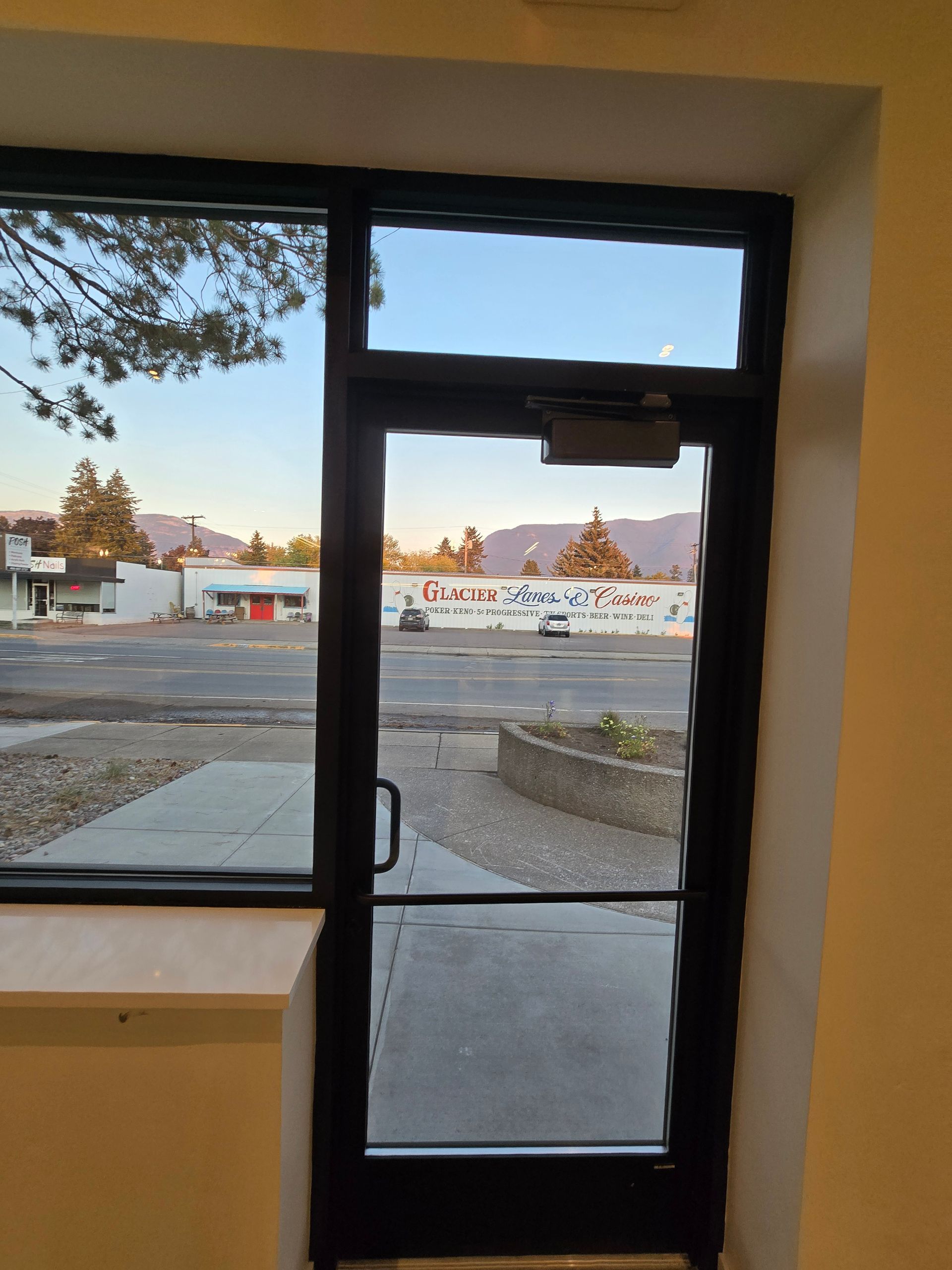A black-framed door and adjacent window look out onto a sunlit parking lot, a painted concrete wall, and distant hills.