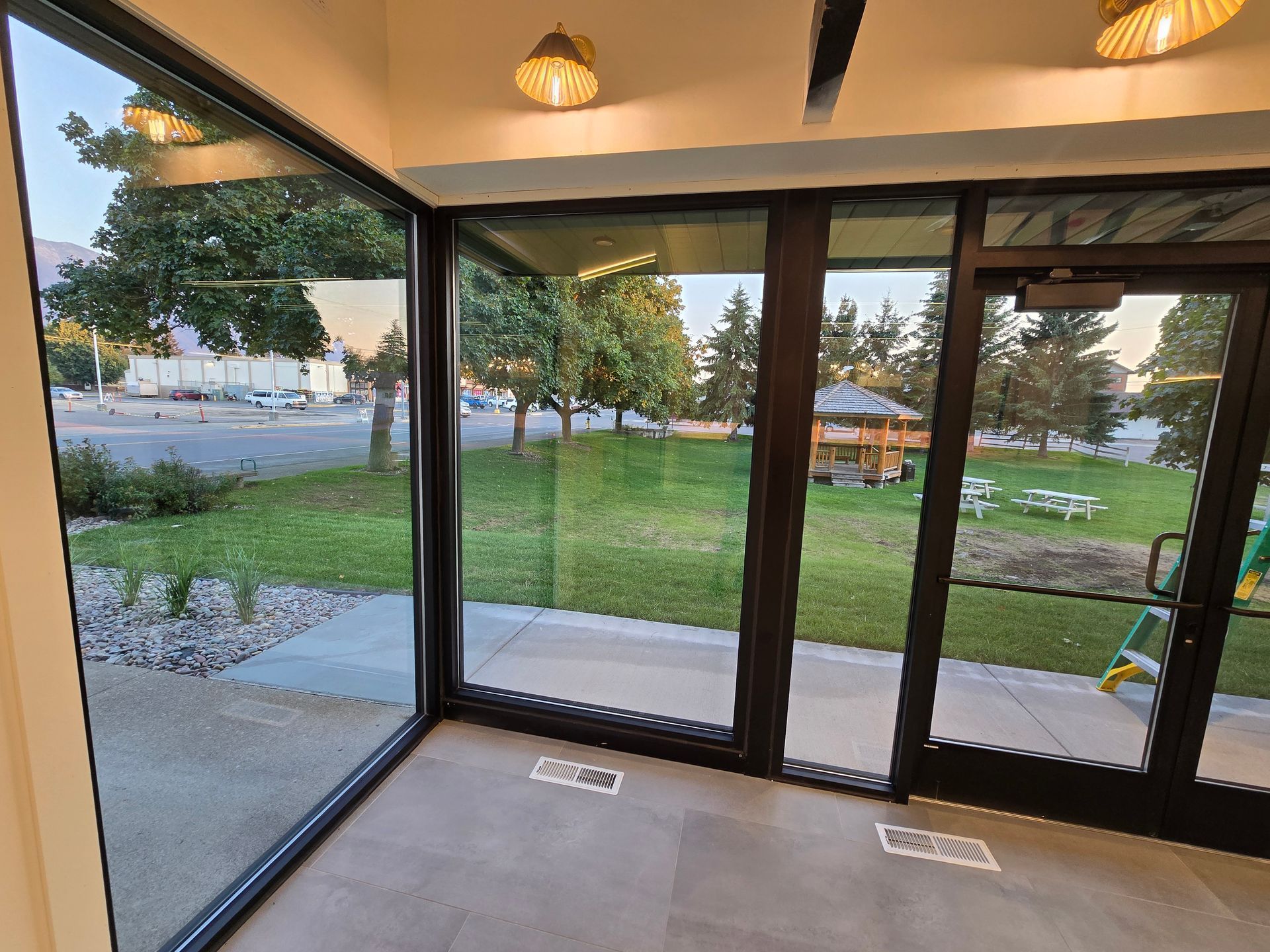 Floor-to-ceiling windows look out from a tiled indoor space onto a grassy park with a gazebo and outdoor seating.