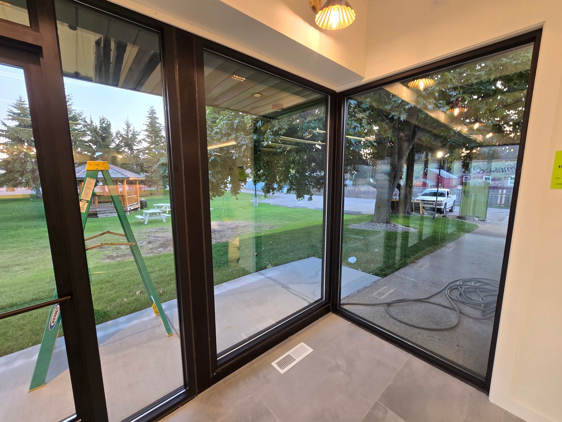 Floor-to-ceiling windows showing a green lawn, trees, and a walkway outside an interior tiled room.