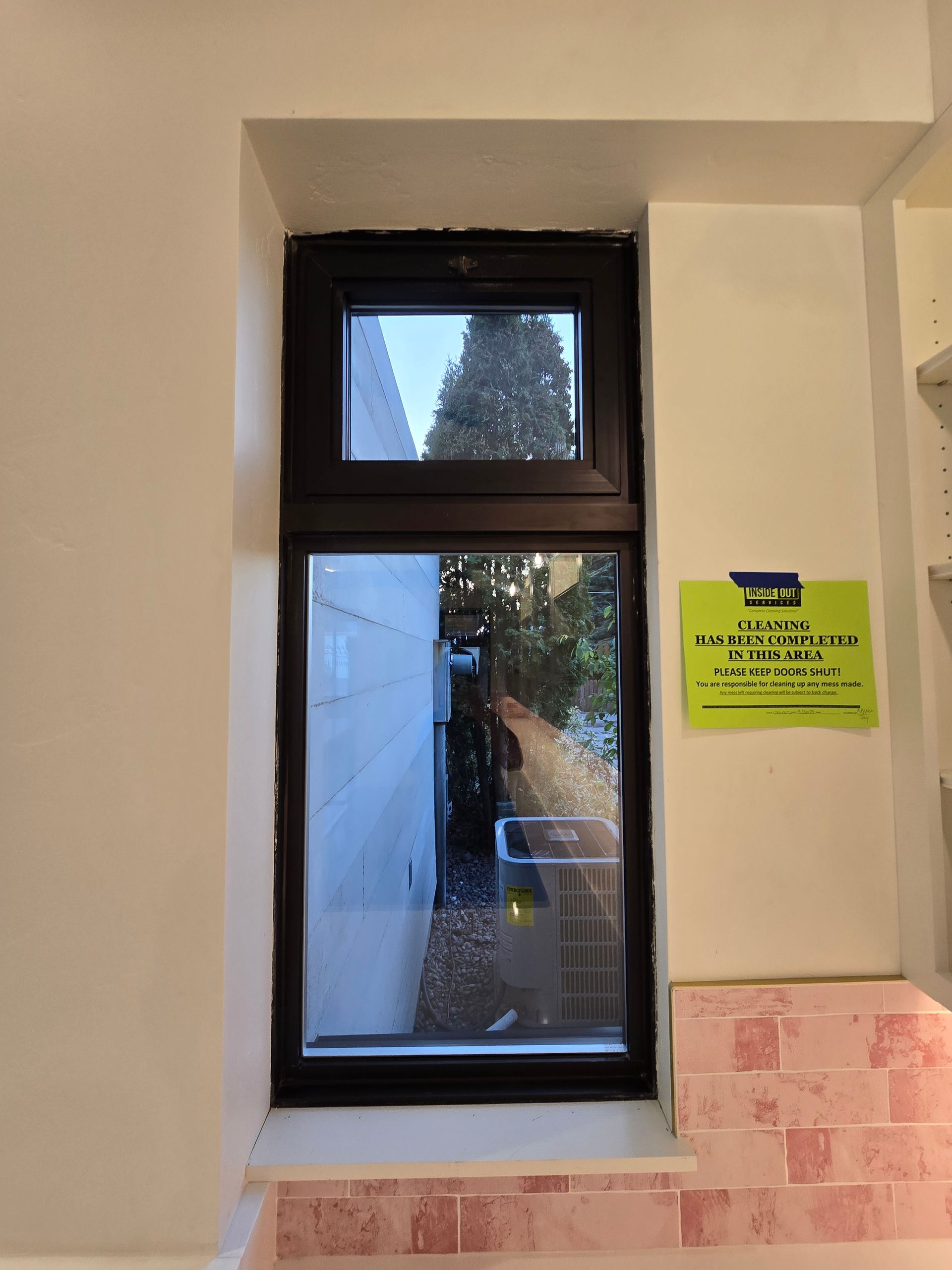 A tall, two-pane dark-framed window overlooking a narrow outdoor space with a bright green notice posted on the adjacent wall.