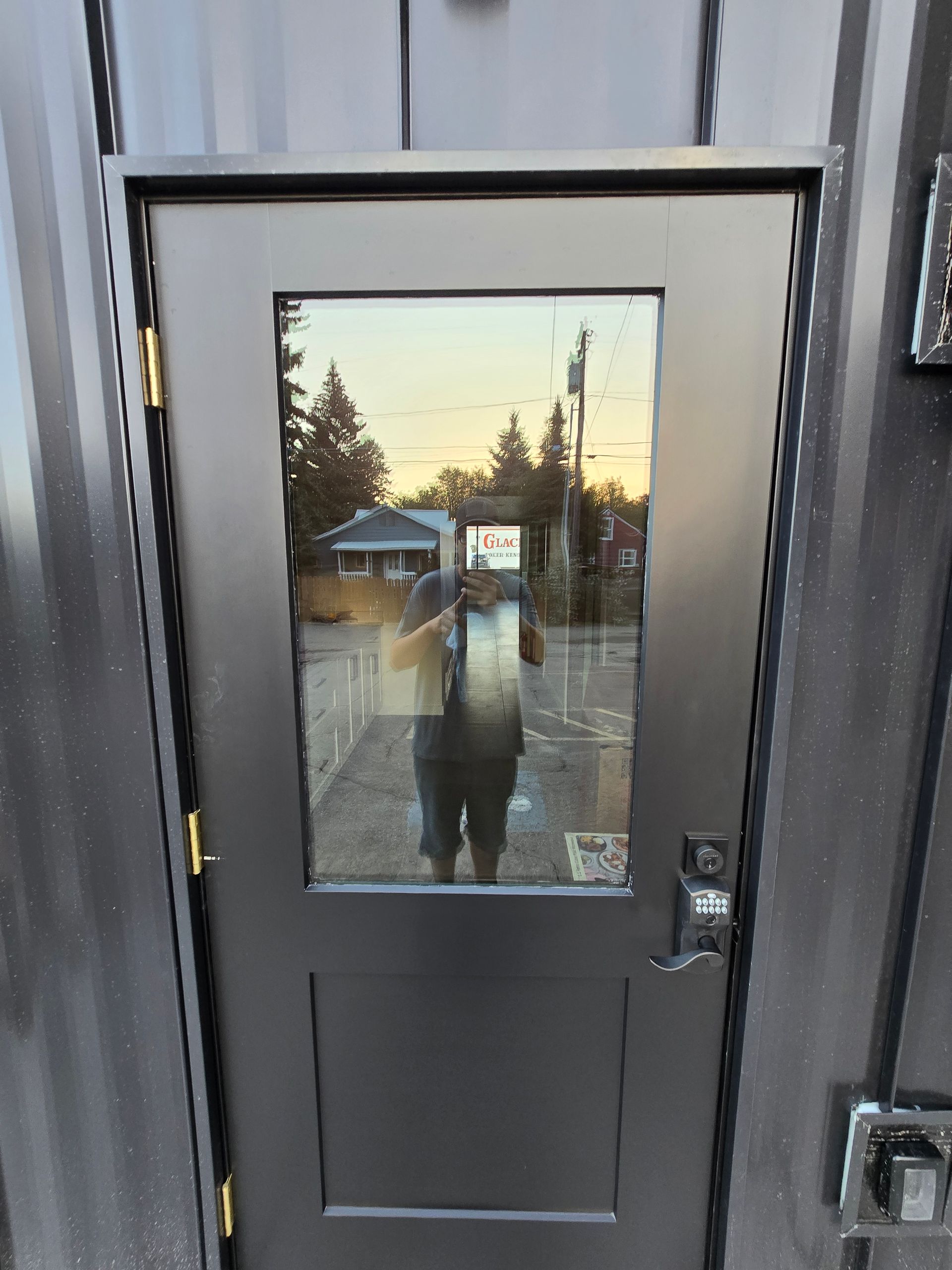 A dark-colored door with a rectangular window reflecting an outdoor scene and a person taking the photo.