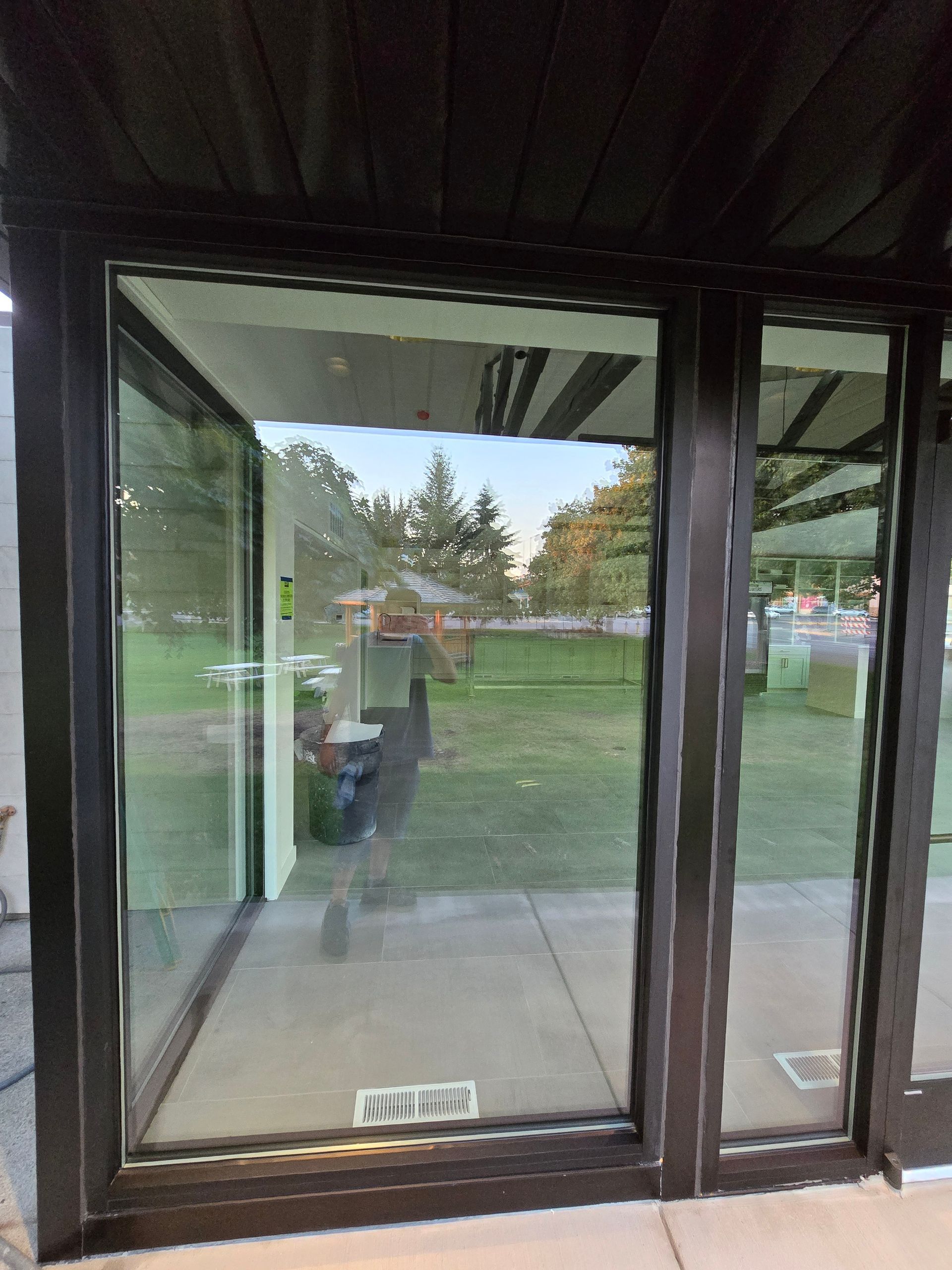 Large glass panels in a dark metal frame reflect a green lawn, trees, and a person walking outdoors.
