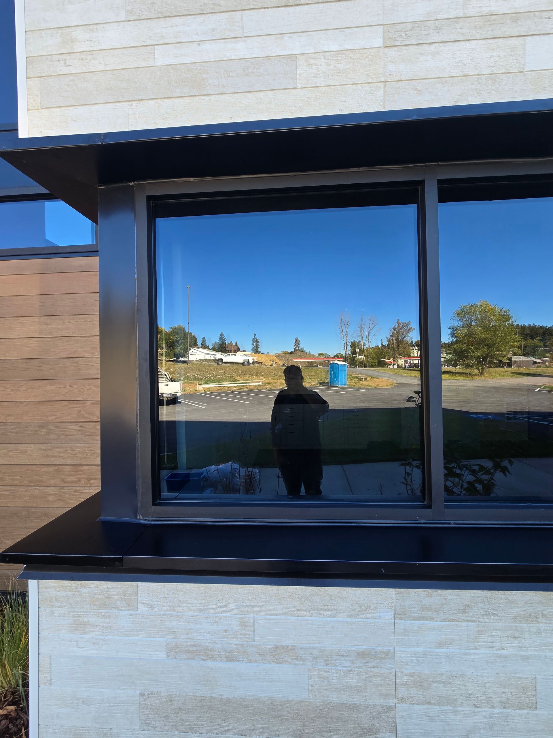 A person stands outside and is reflected in the large, tinted glass window of a building with light stone siding.