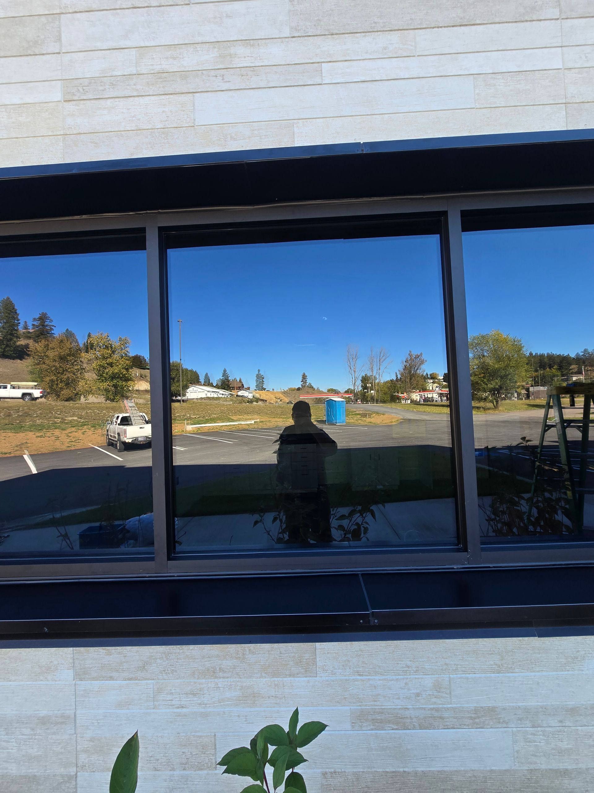 A person is reflected in a large, modern office window showing a bright, sunny outdoor parking lot and landscape.