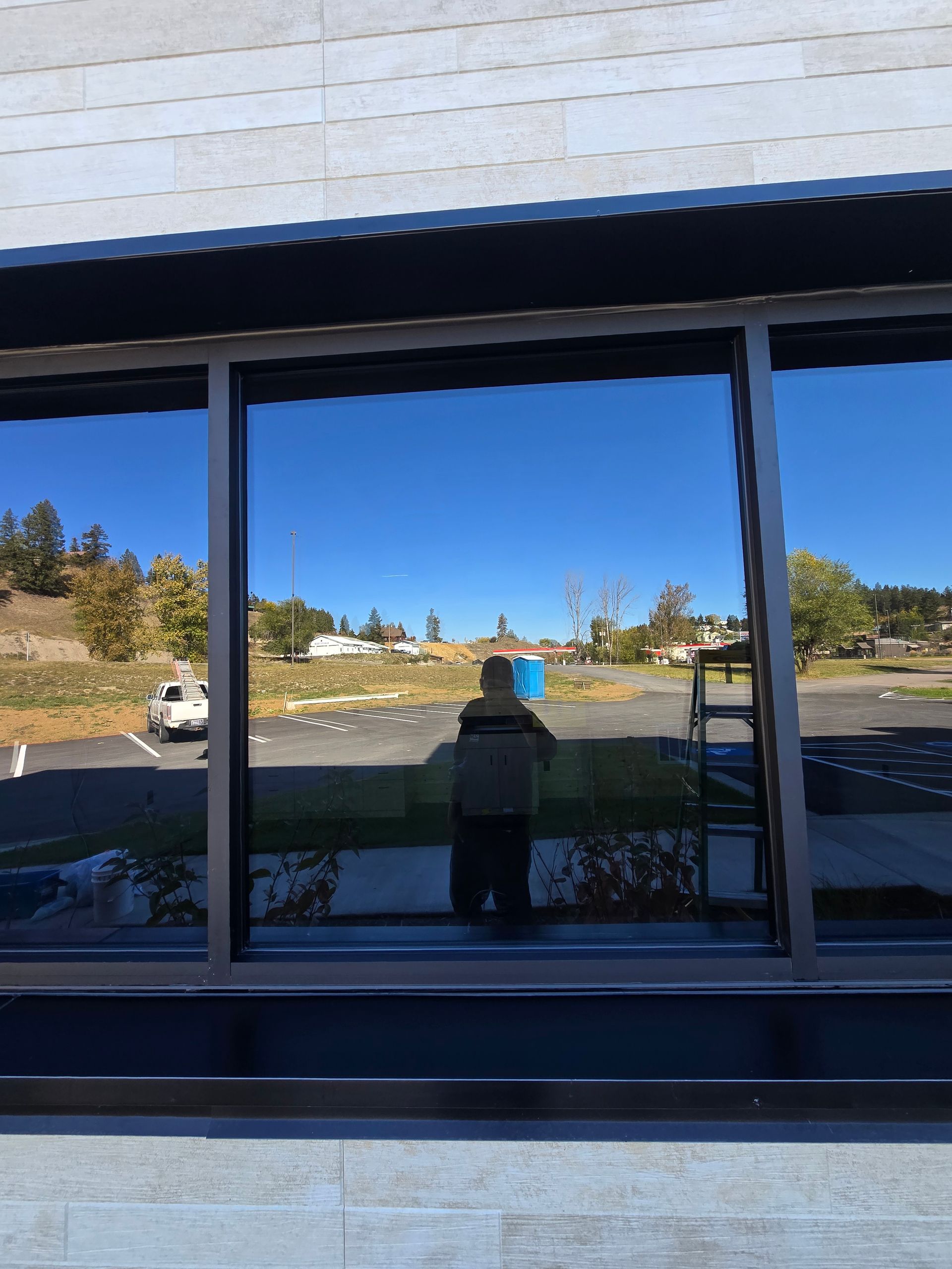 A person stands in front of a reflective glass window, which mirrors an outdoor parking lot, trees, and a clear blue sky.