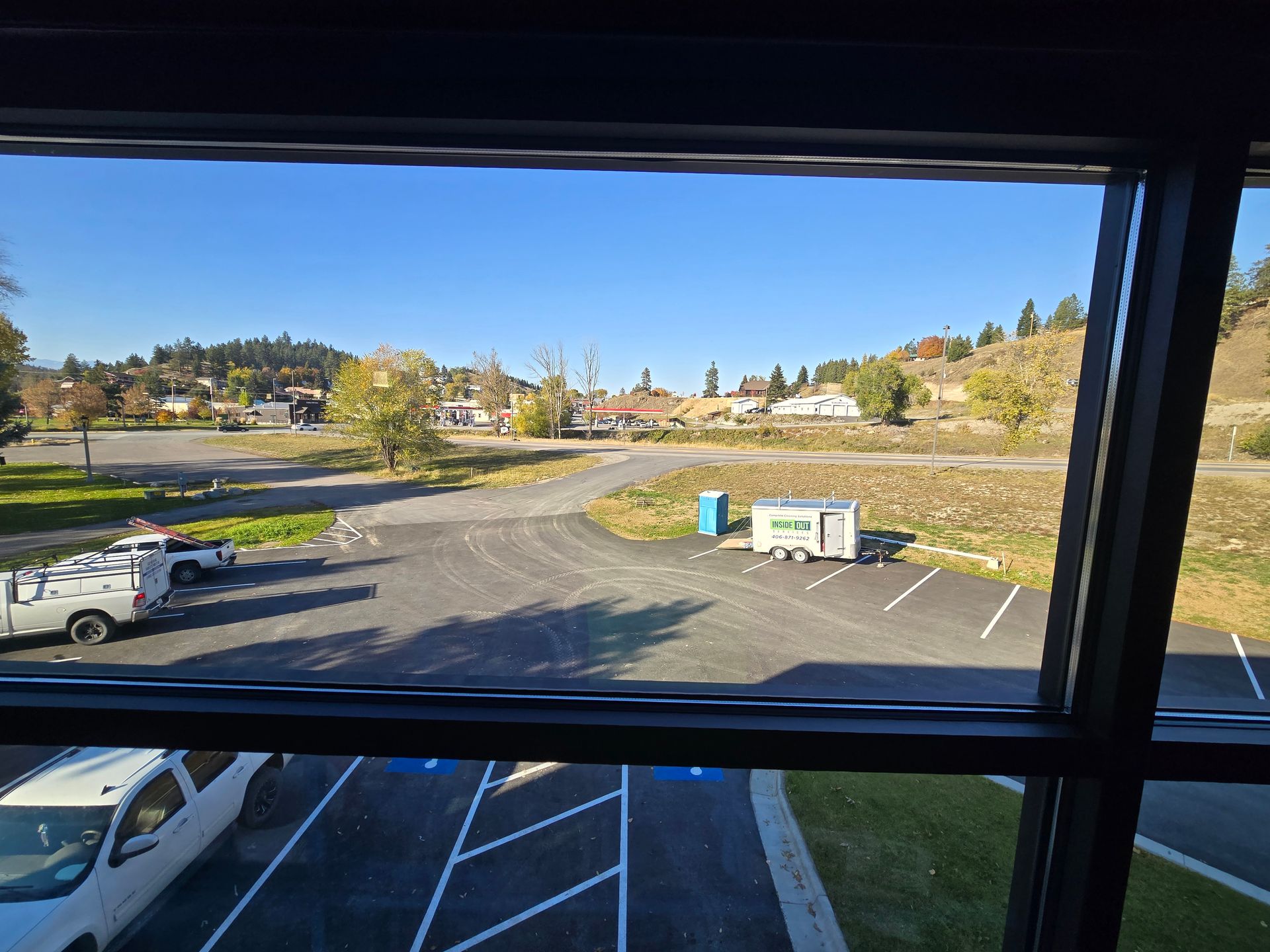 A parking lot viewed through a window, with a white truck, a portable toilet, and a small trailer on a sunny day.