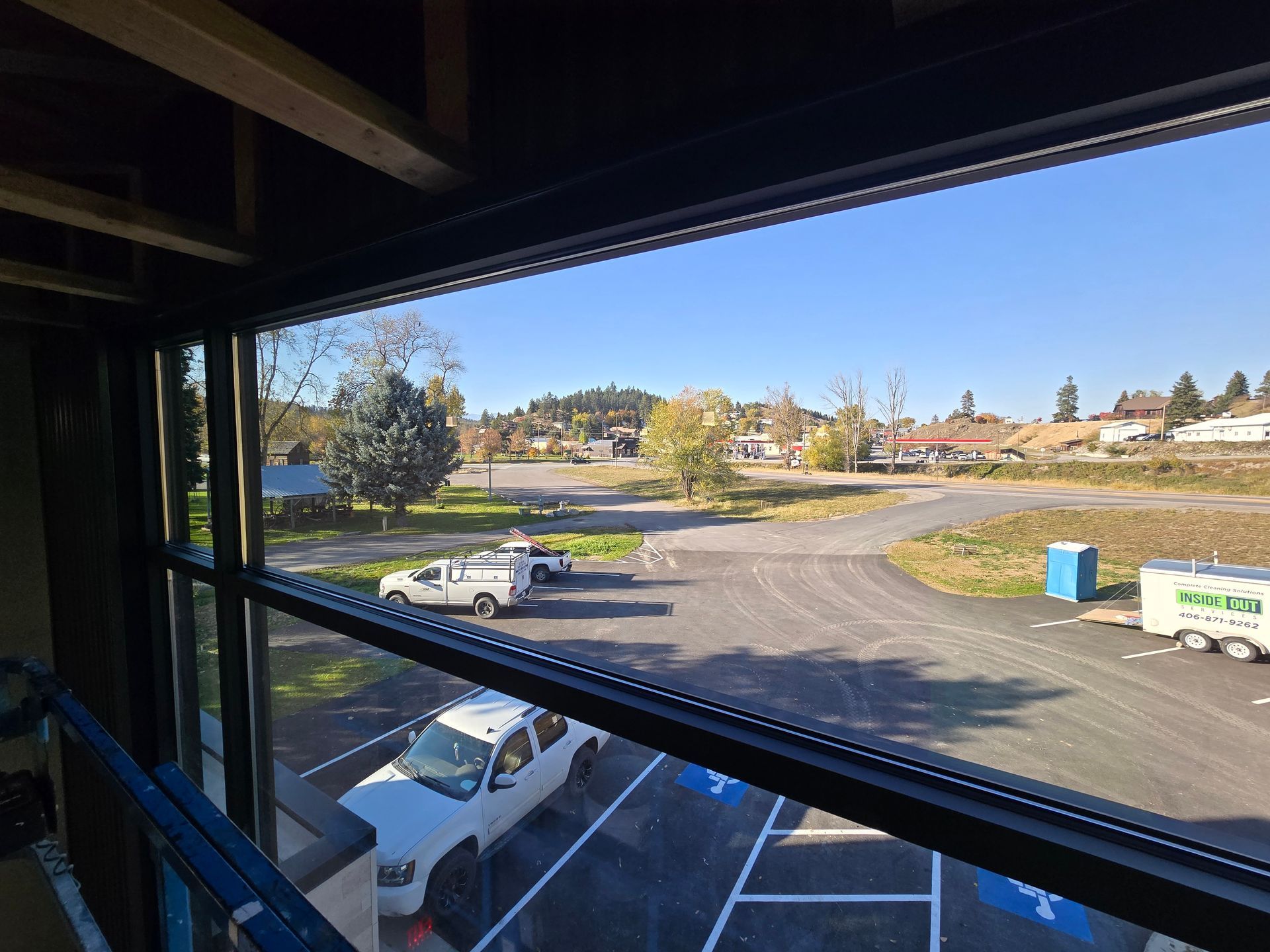 View from an indoor construction site looking out over a parking lot, a grassy field, and trees under a clear blue sky.