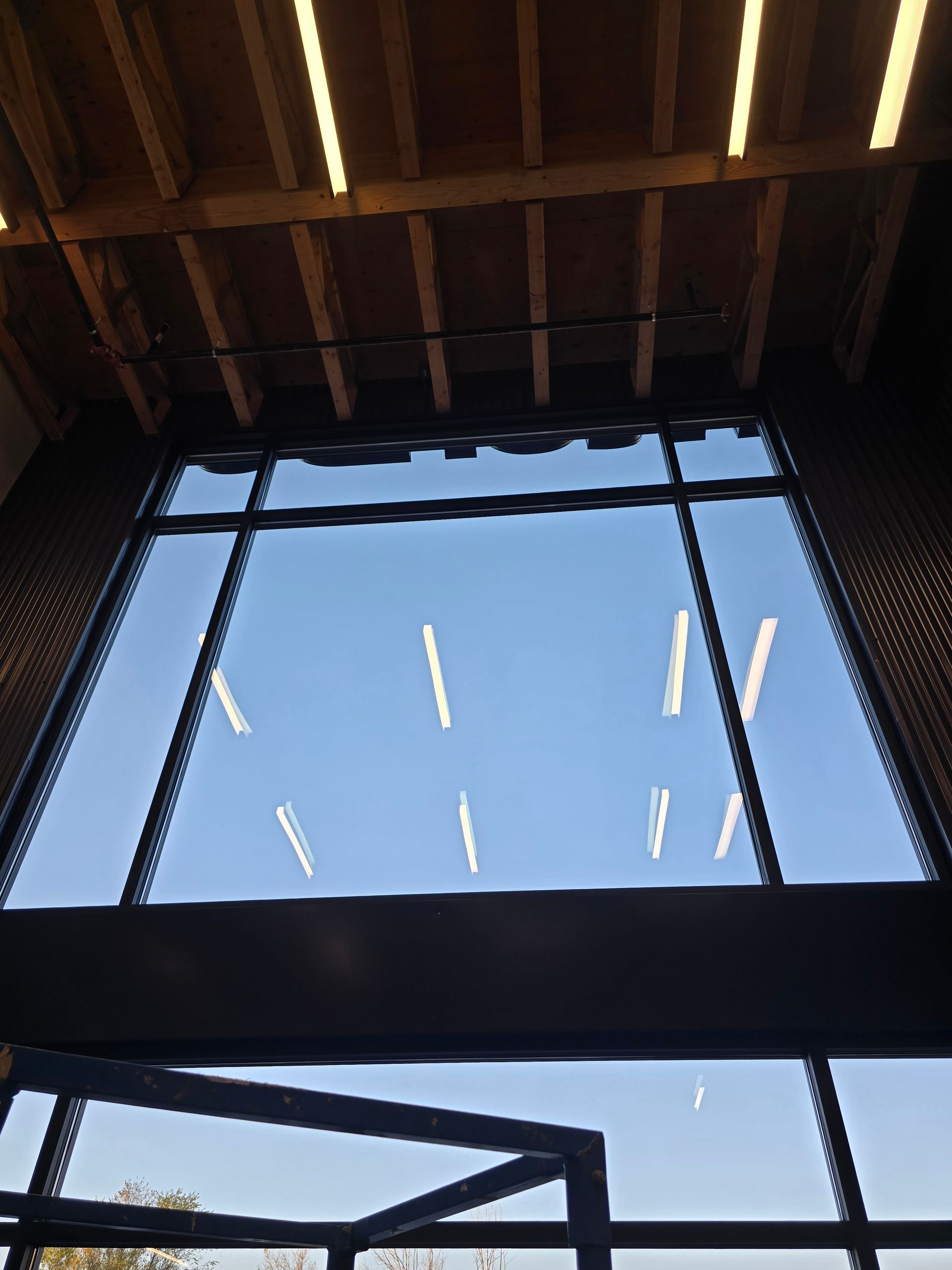 Large floor-to-ceiling windows showing a bright blue sky, with interior ceiling beams and reflections of light fixtures.