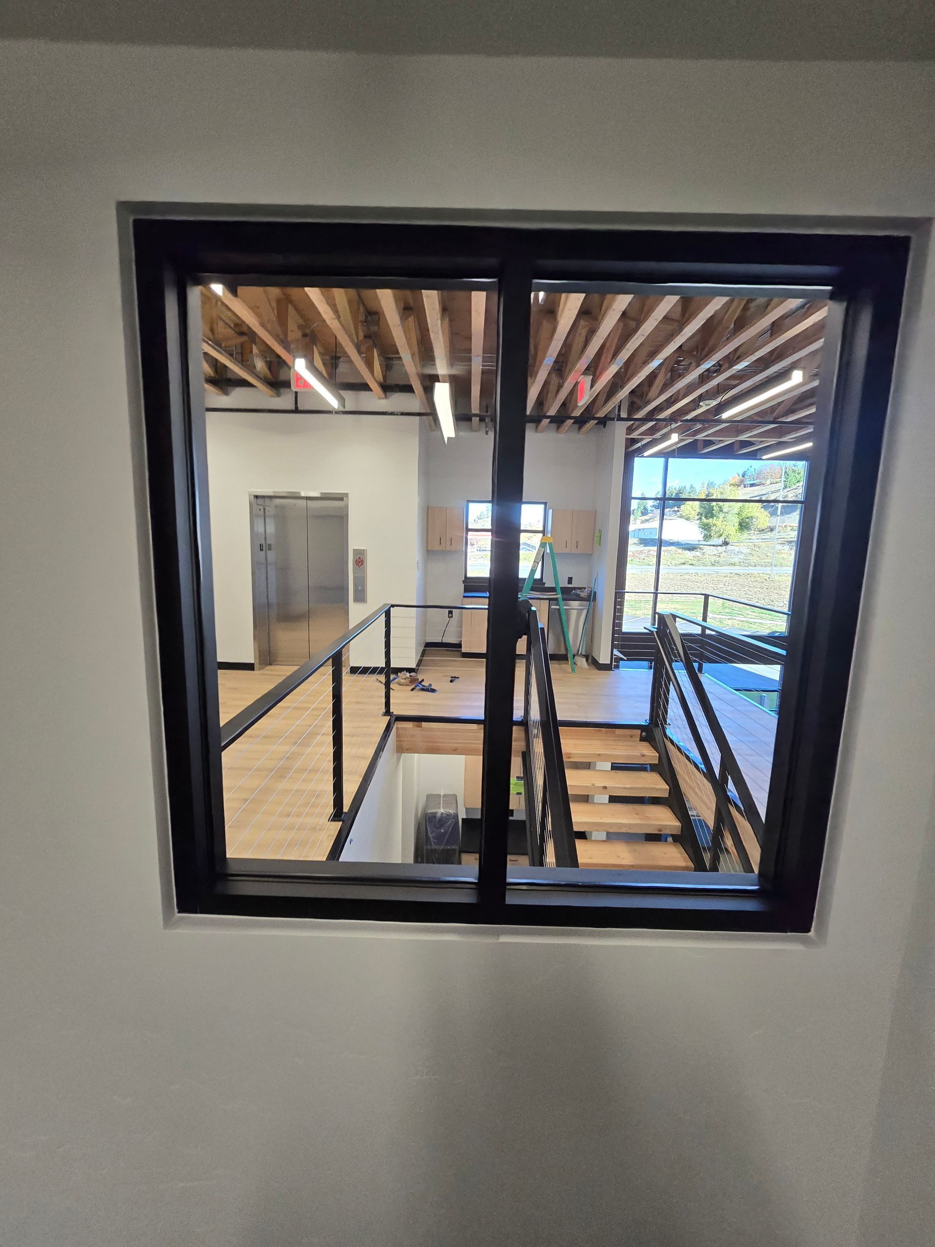 View through a black-framed window into a building interior featuring an open stairwell, light wood floors, and railings.