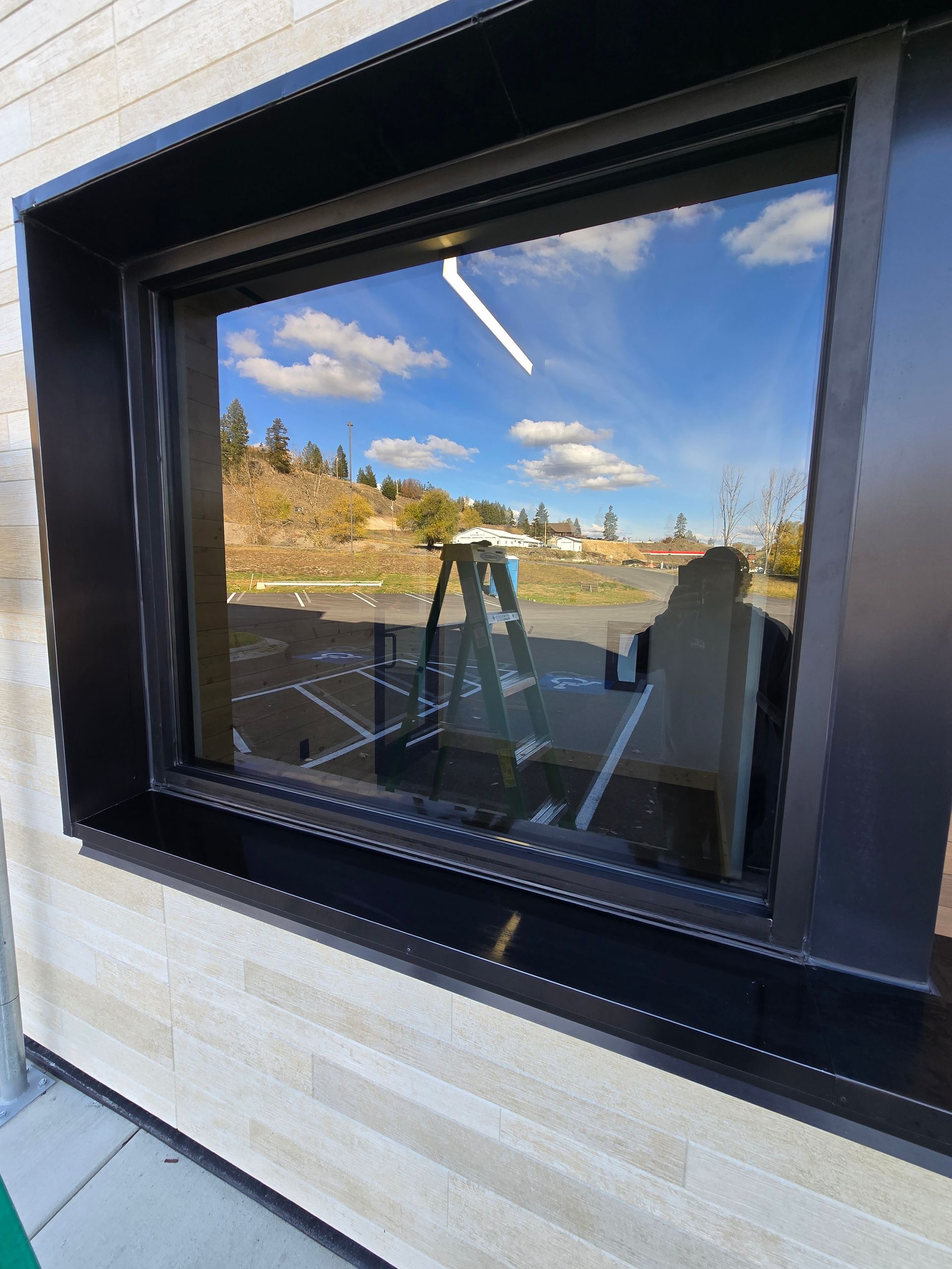 A black-framed window reflecting a blue sky, trees, a ladder, and a parking lot on a building with light stone siding.