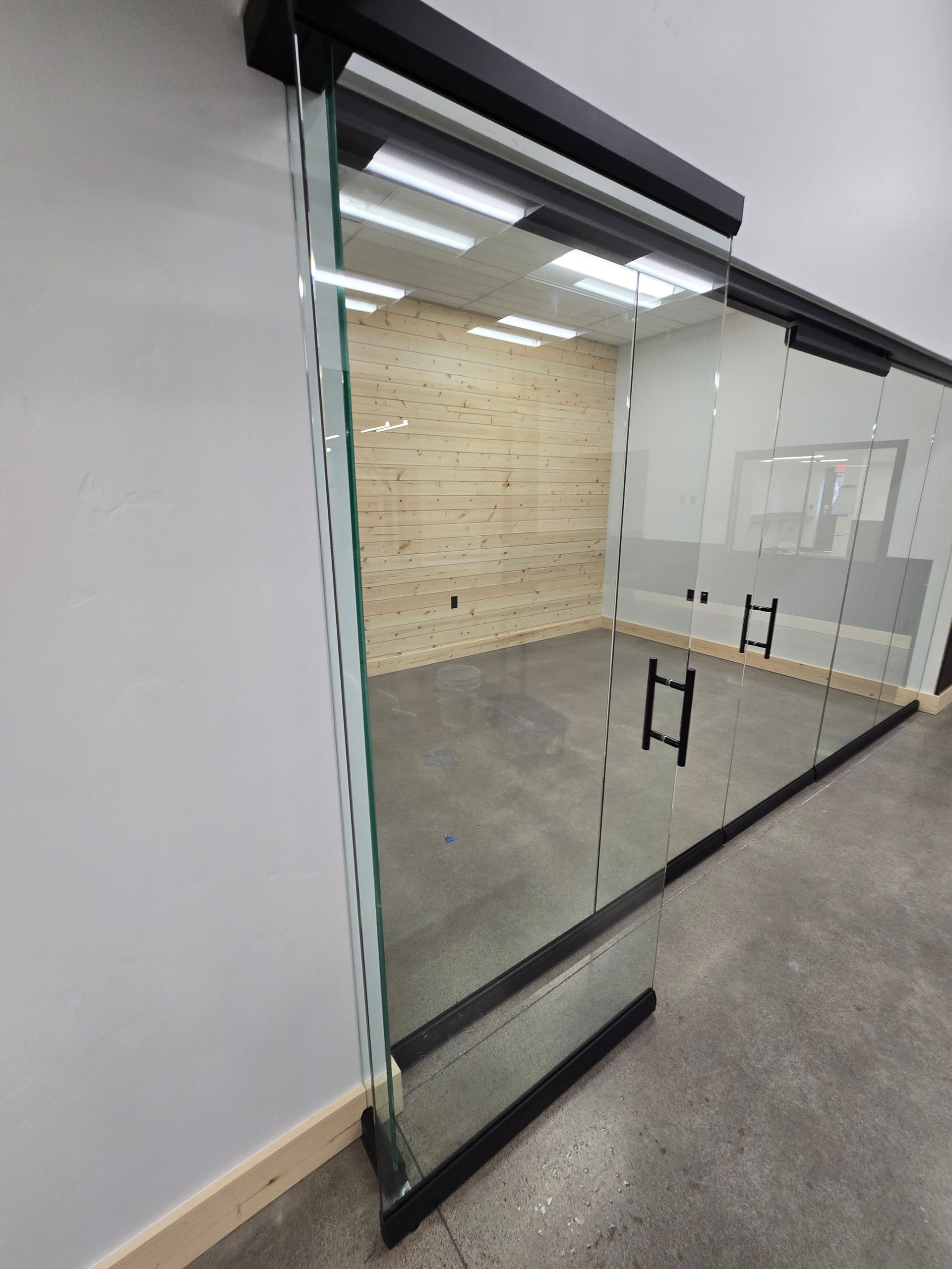 A glass wall partition with a black frame and door handles, enclosing a room with a light wood-paneled wall.