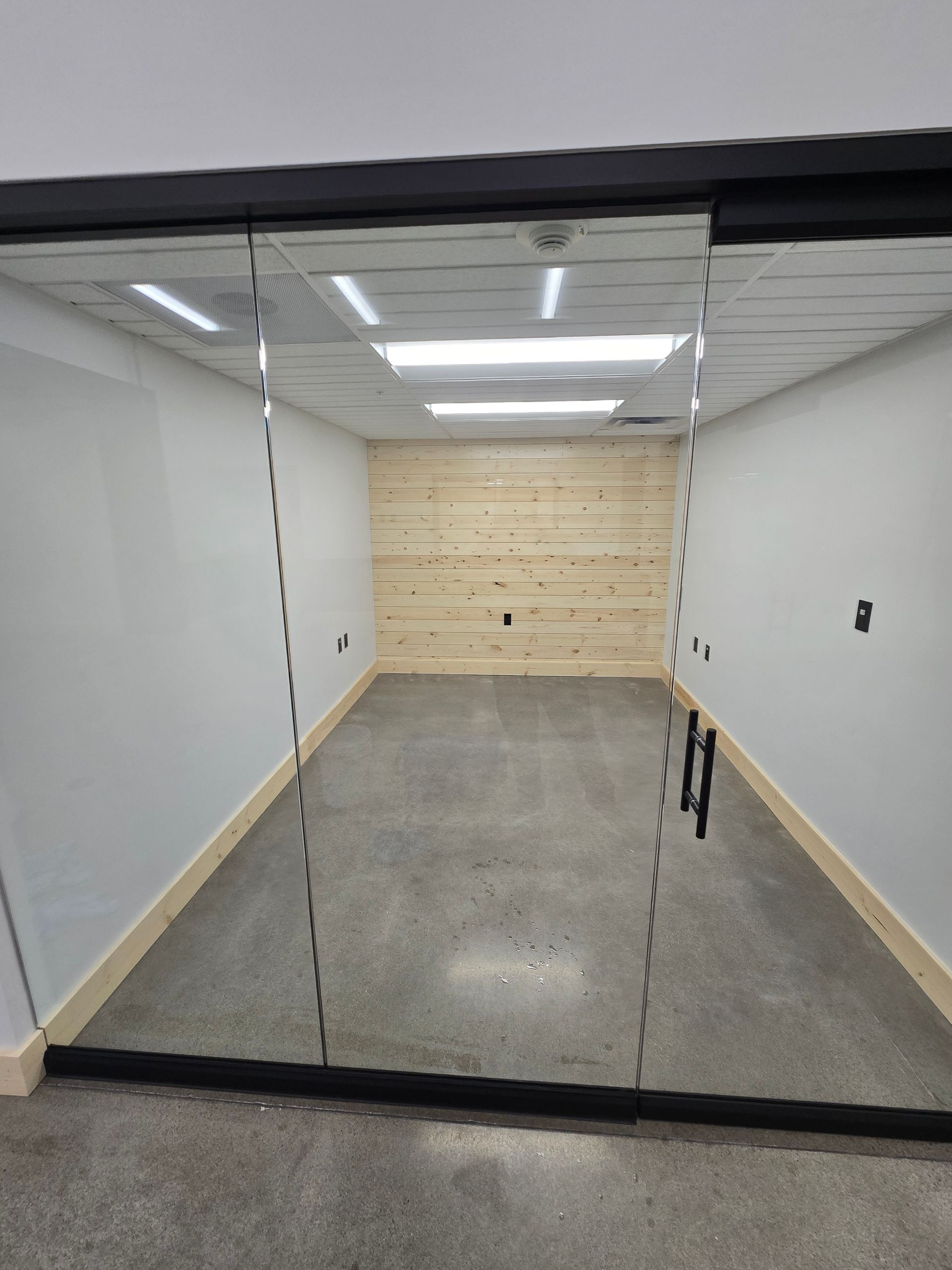 A modern office meeting room with glass walls, a gray concrete floor, white walls, and a wood-paneled back wall.