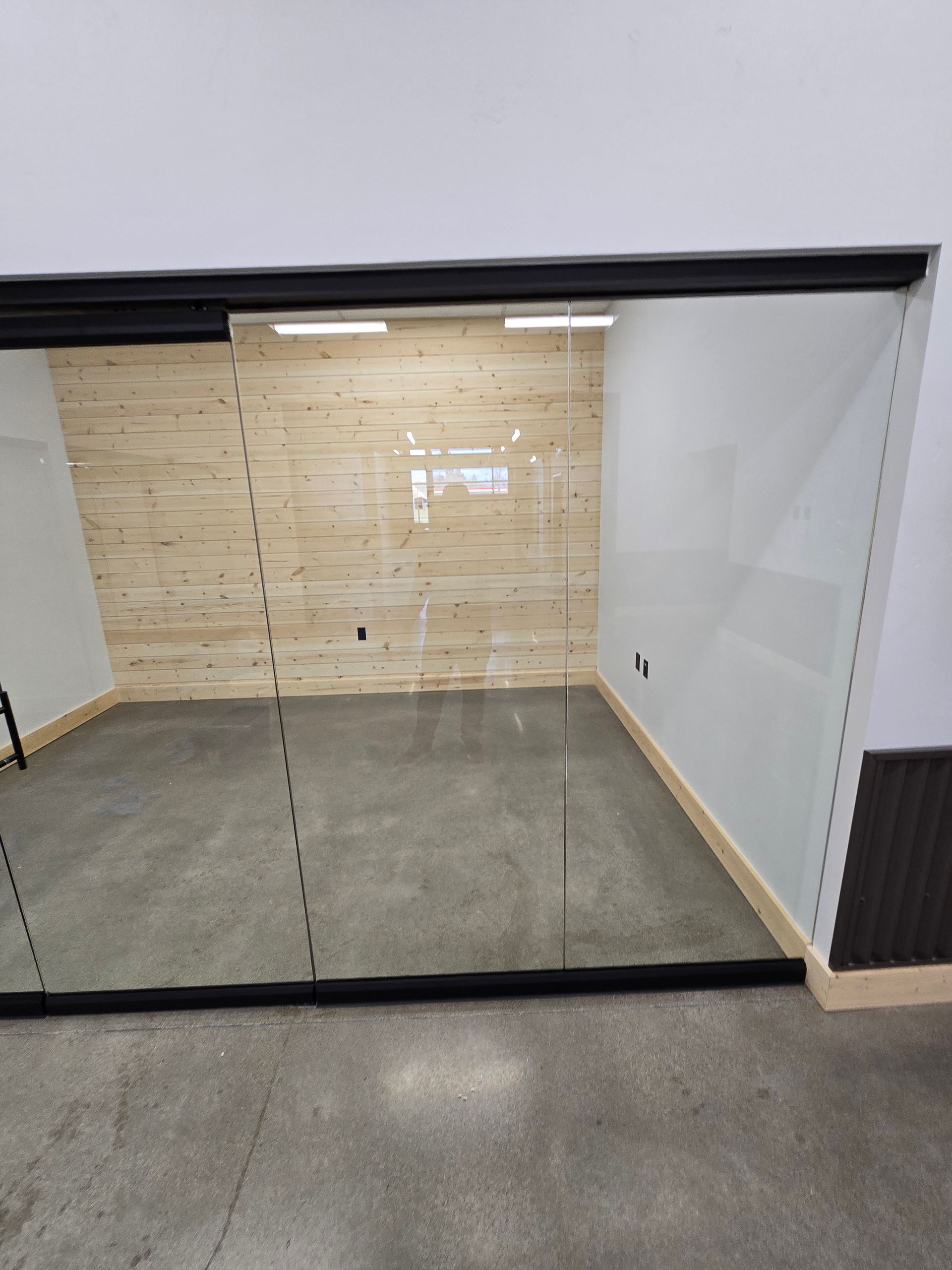 A glass-walled office with a light wood-paneled back wall and gray concrete flooring, viewed from a white-walled exterior.